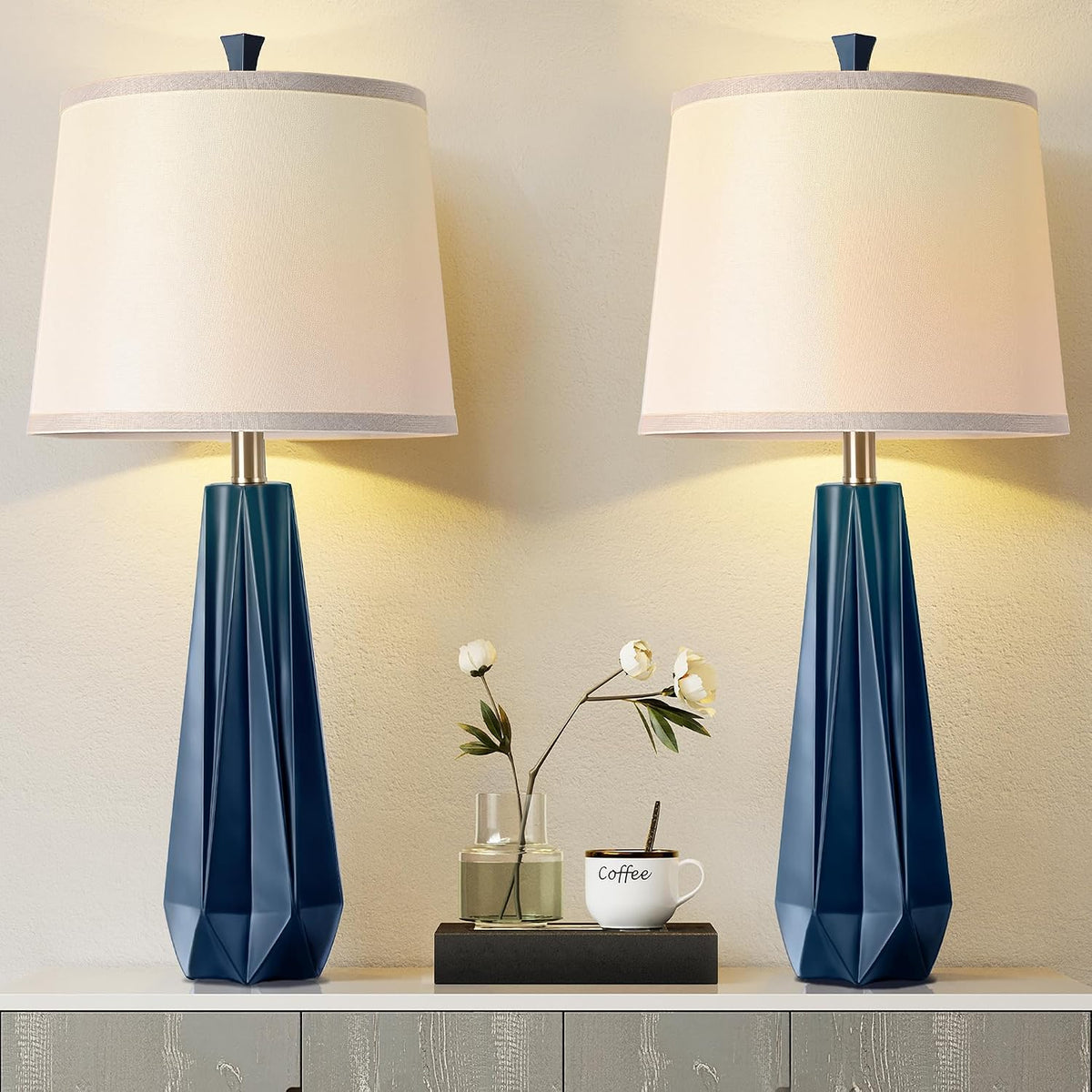 Front view of two deep-blue geometric table lamps with beige linen shades positioned with a vase and coffee cup between them.