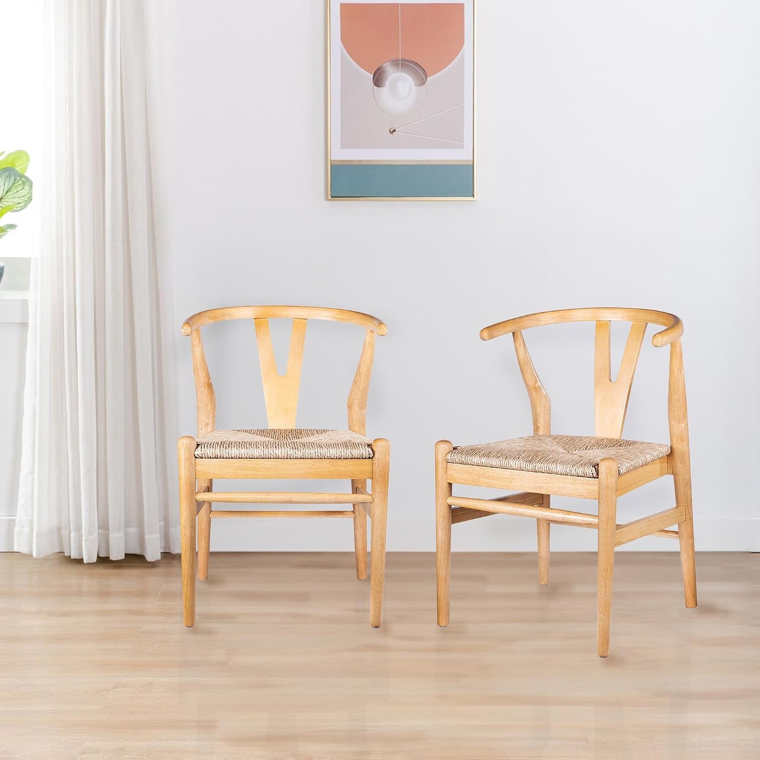 Two natural-finish wishbone chairs with woven seats on a light wood floor against a pale wall with framed art and a curtain