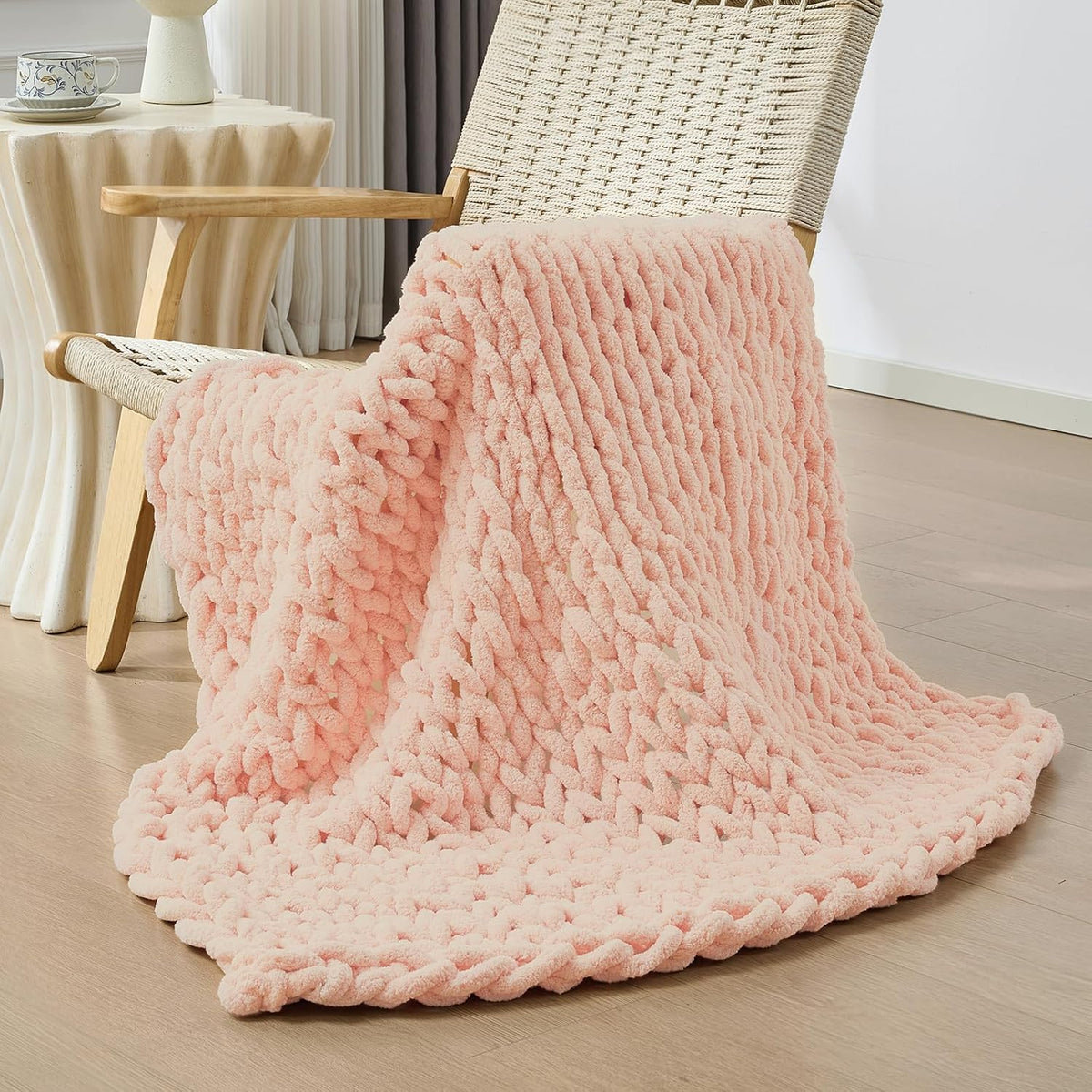 Soft blush-pink chunky knit throw draped over a woven lounge chair, pooling onto a light wood floor.