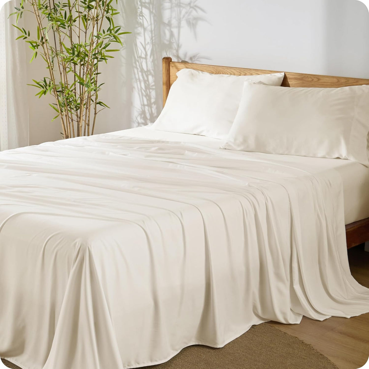 Bed made with ivory silky sheets and two pillows against a wooden headboard and potted bamboo