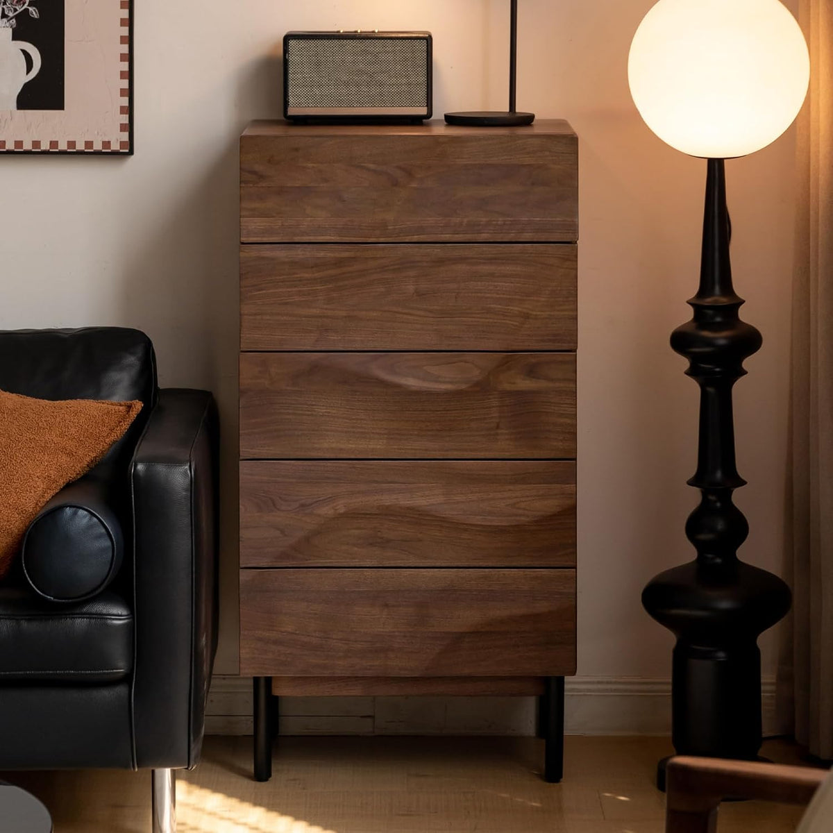 Tall narrow chest photographed in a living area beside a sofa and lamp, showing its slim profile and raised legs.
