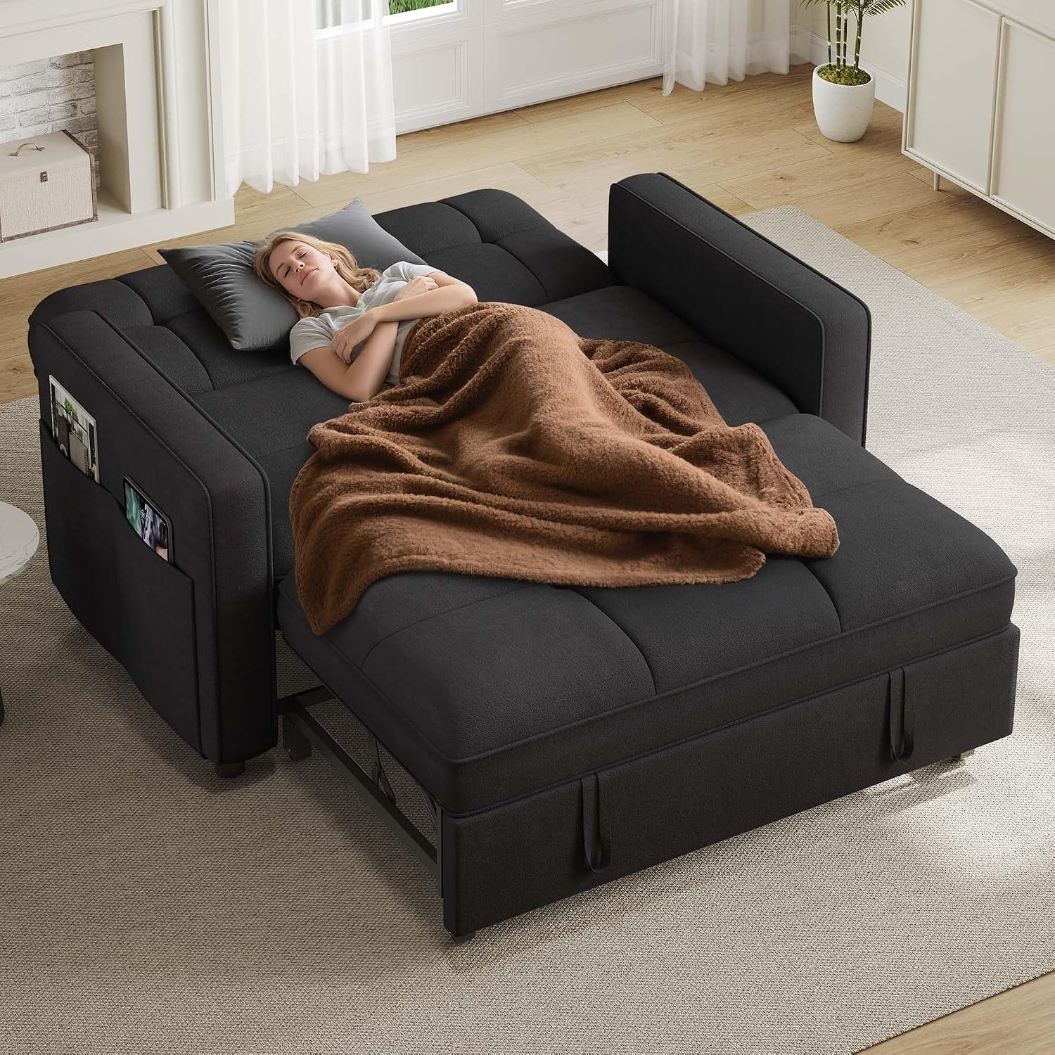 Top-down view of a dark gray futon sofa bed opened flat with a brown blanket and a pillow, set in a sunlit living room.
