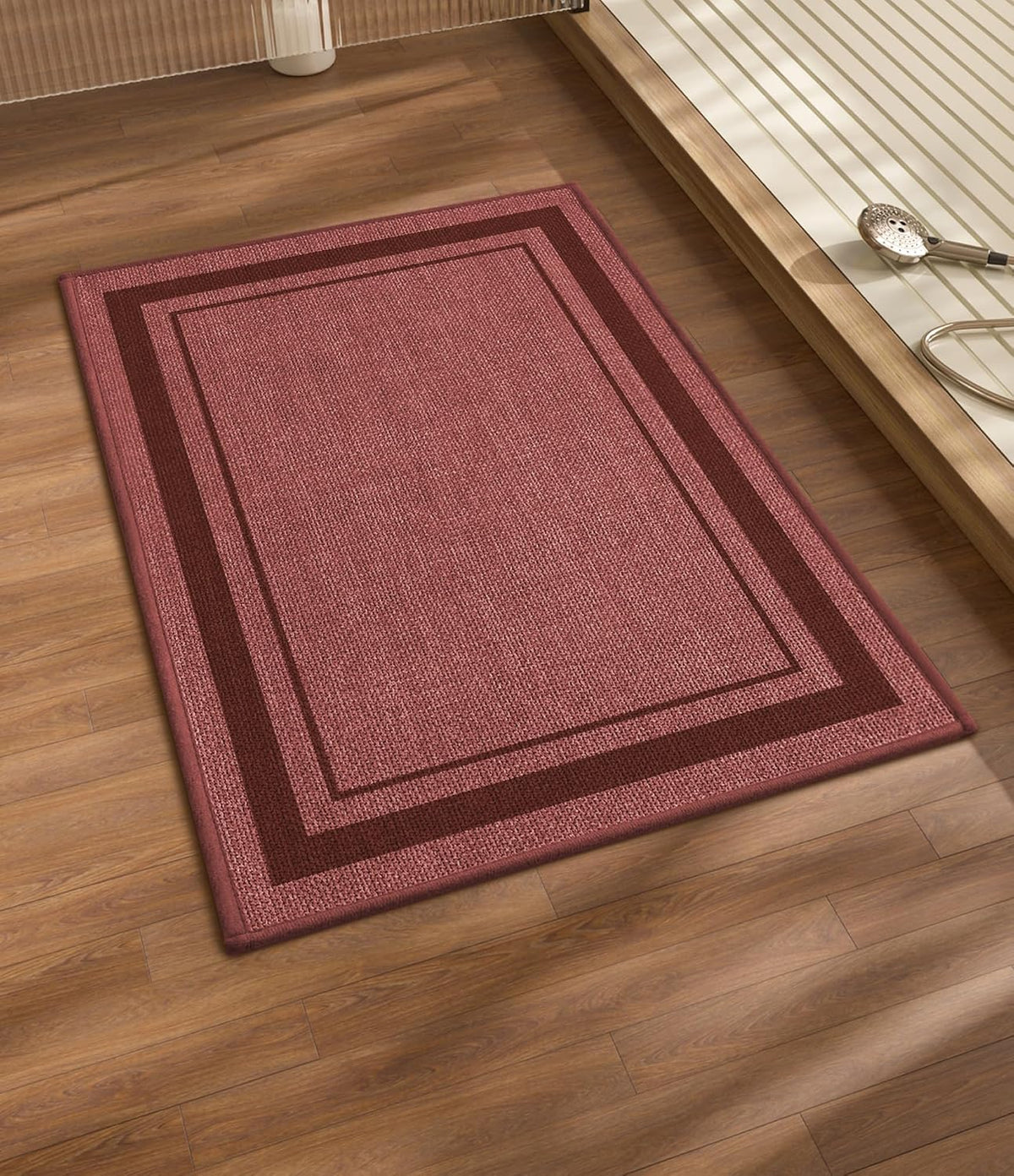 Muted rose rectangular bath rug with darker double border on a wooden floor near a tub