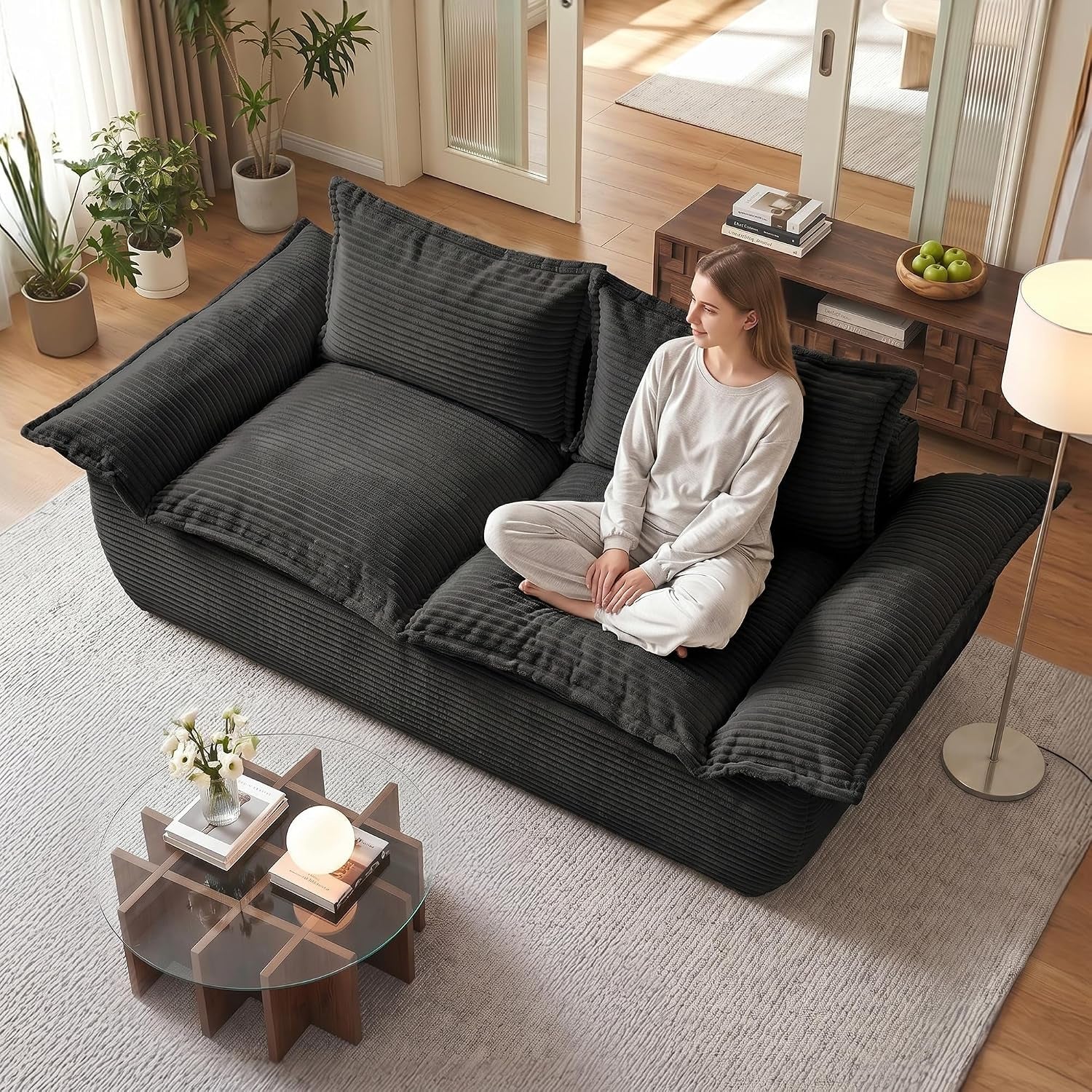 Deep charcoal corduroy two-seater sofa with oversized cushions; woman sitting cross-legged, coffee table, plants and lamp