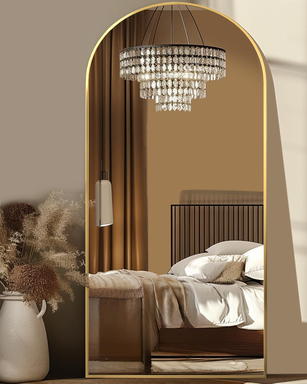 Gold arched mirror with thin frame reflecting a crystal chandelier and a styled bedroom scene with bed and drapery.