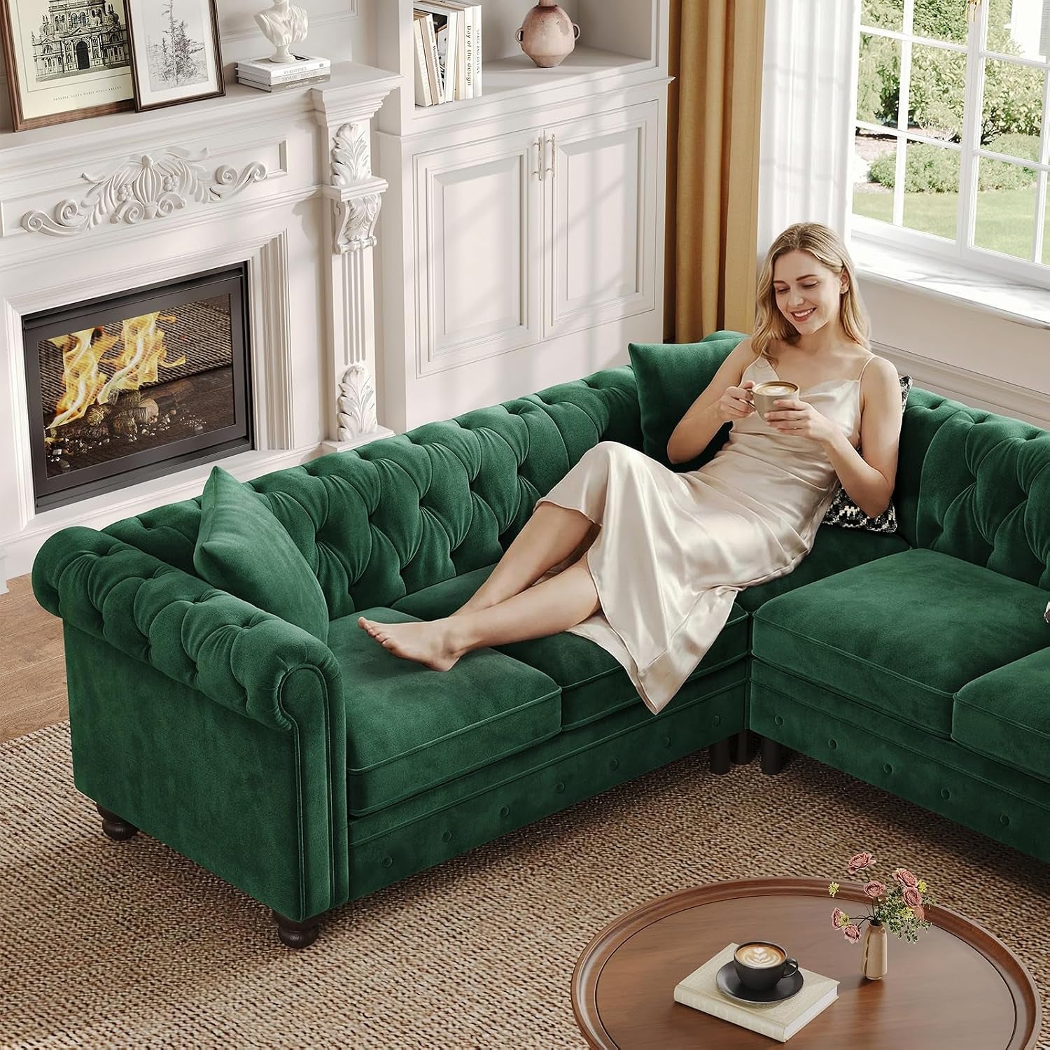 Woman reclining on the green velvet L-shaped sectional beside a lit fireplace, holding a cup