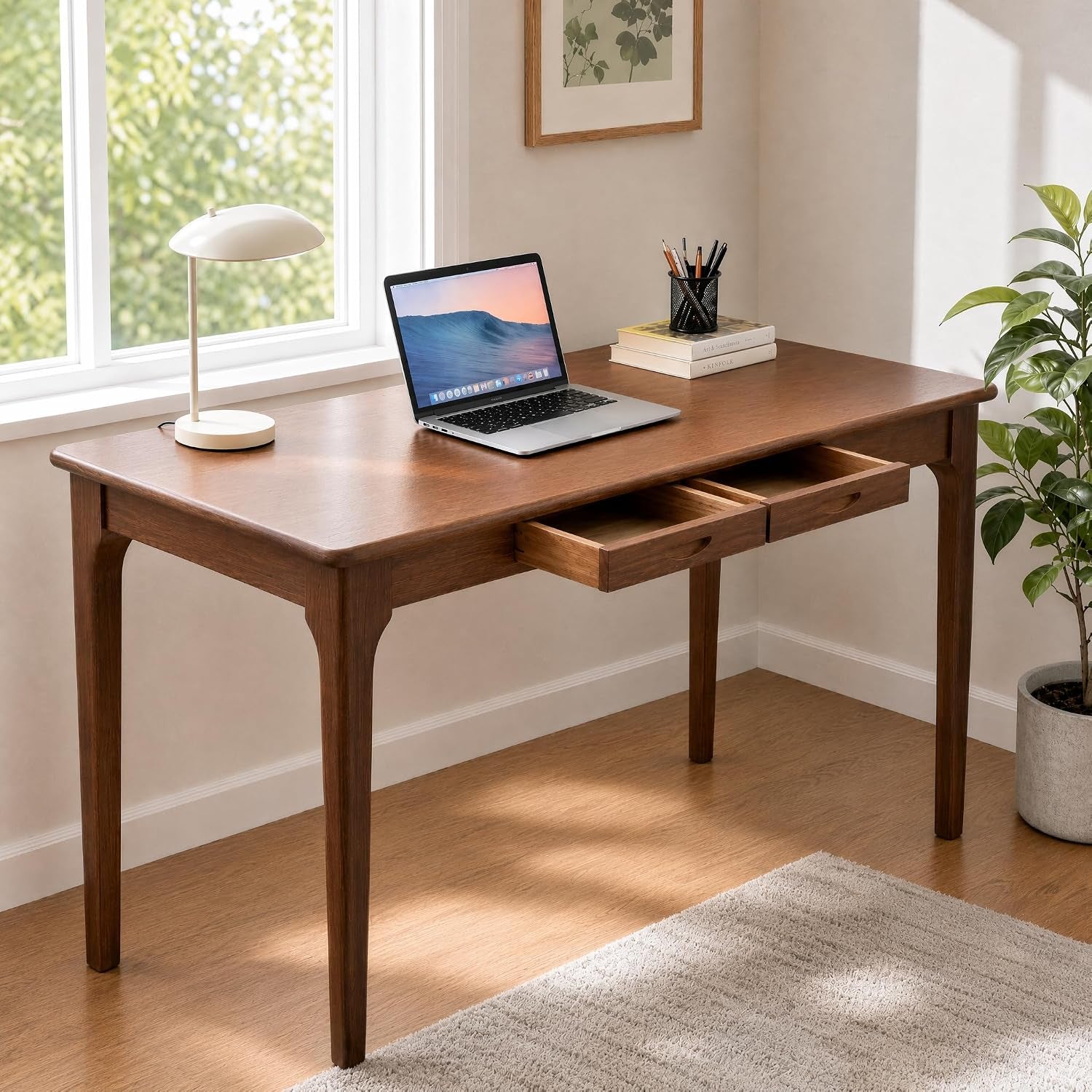 Three-quarter view of the desk by a bright window with a laptop, lamp and one drawer pulled out.