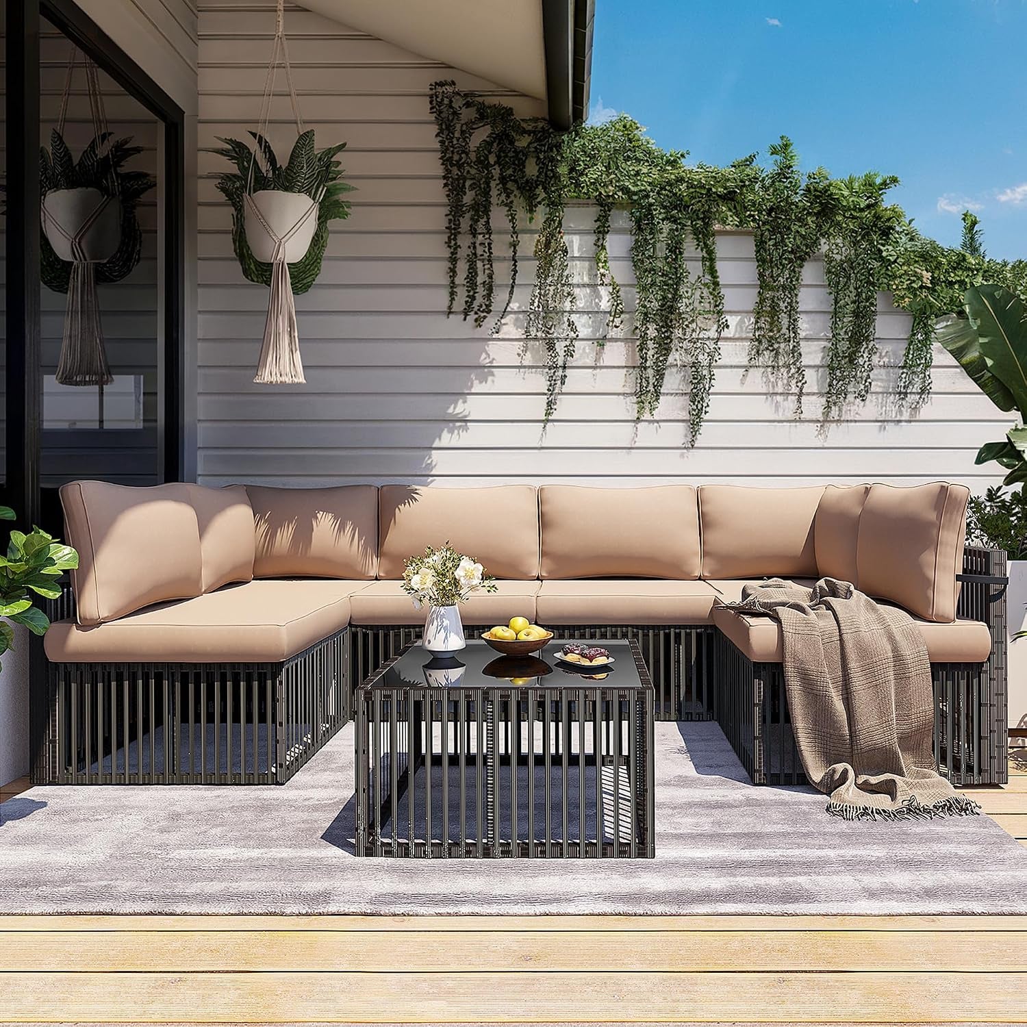 Outdoor L-shaped sectional with tan cushions and black slatted metal coffee table under hanging plants on a deck.