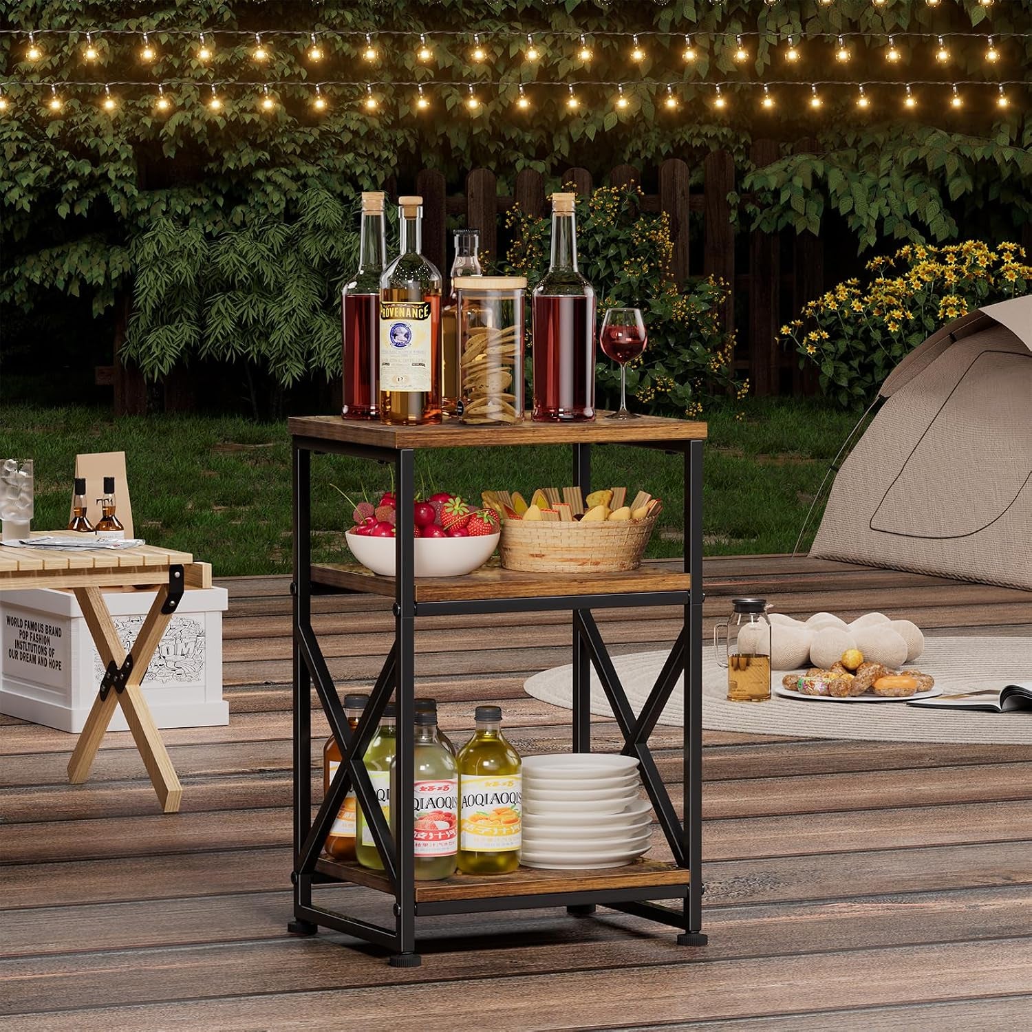 Three-tier rustic side table on a wooden deck holding bottles, bowls and plates under string lights.