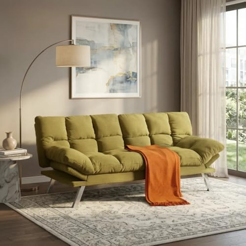 Angled view of an olive-green futon sofa with chrome legs and an orange throw across the seat in a sunlit room.