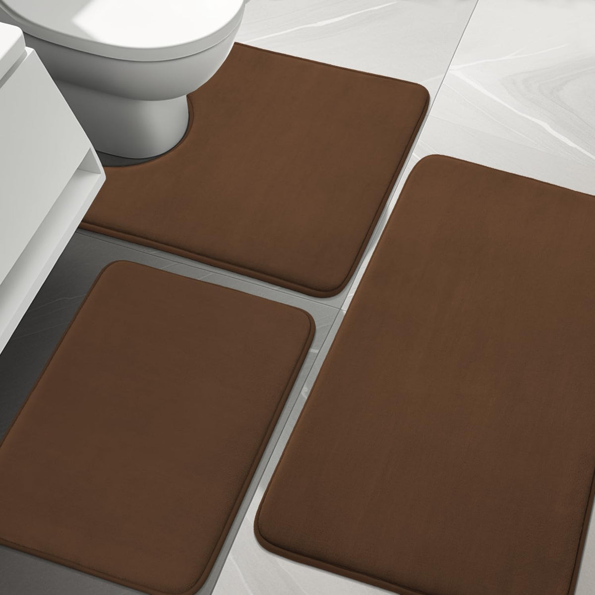 Close overhead crop of three brown bath mats arranged on pale tile near a white toilet base.