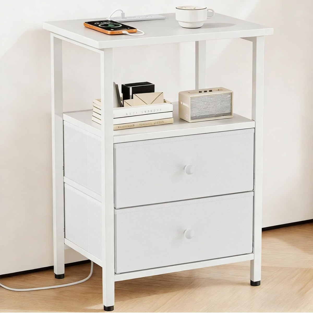 Front three-quarter view of a white nightstand with white metal frame, two fabric drawers and shelf holding books and a