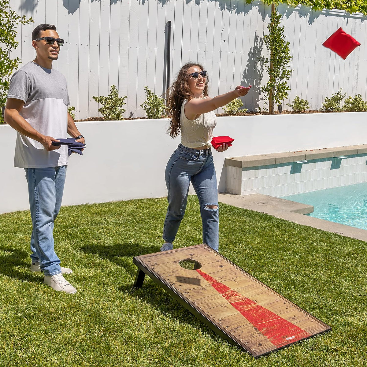 Woman tossing a red cornhole bean bag beside a pool with the board set up on the lawn