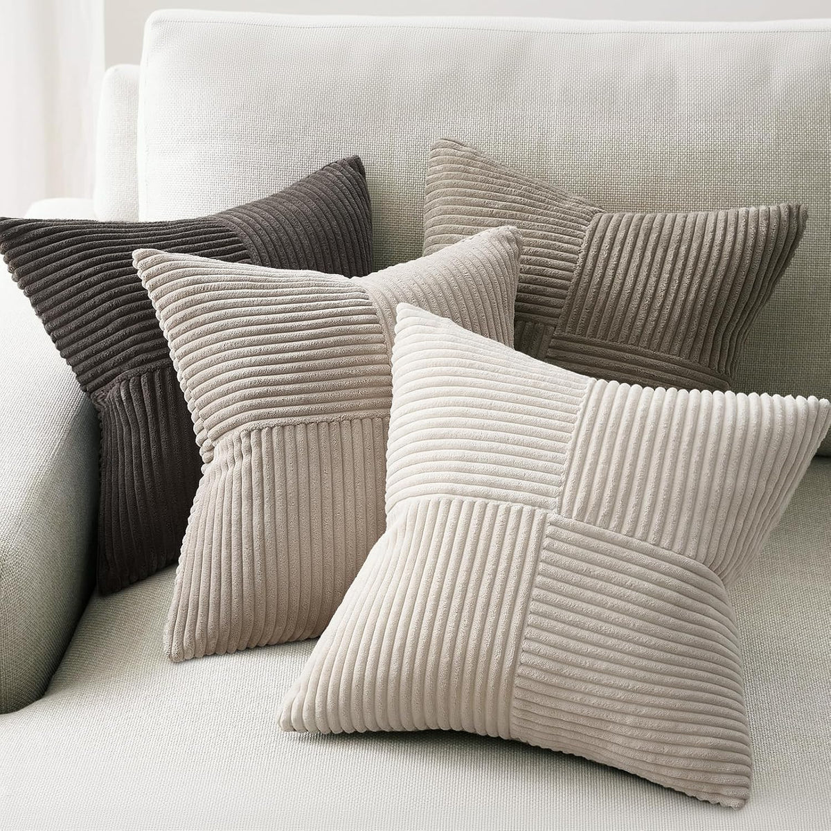 Four ribbed velvet pillow covers in neutral tones—dark charcoal, taupe, light beige, and stone—stacked on a light sofa.