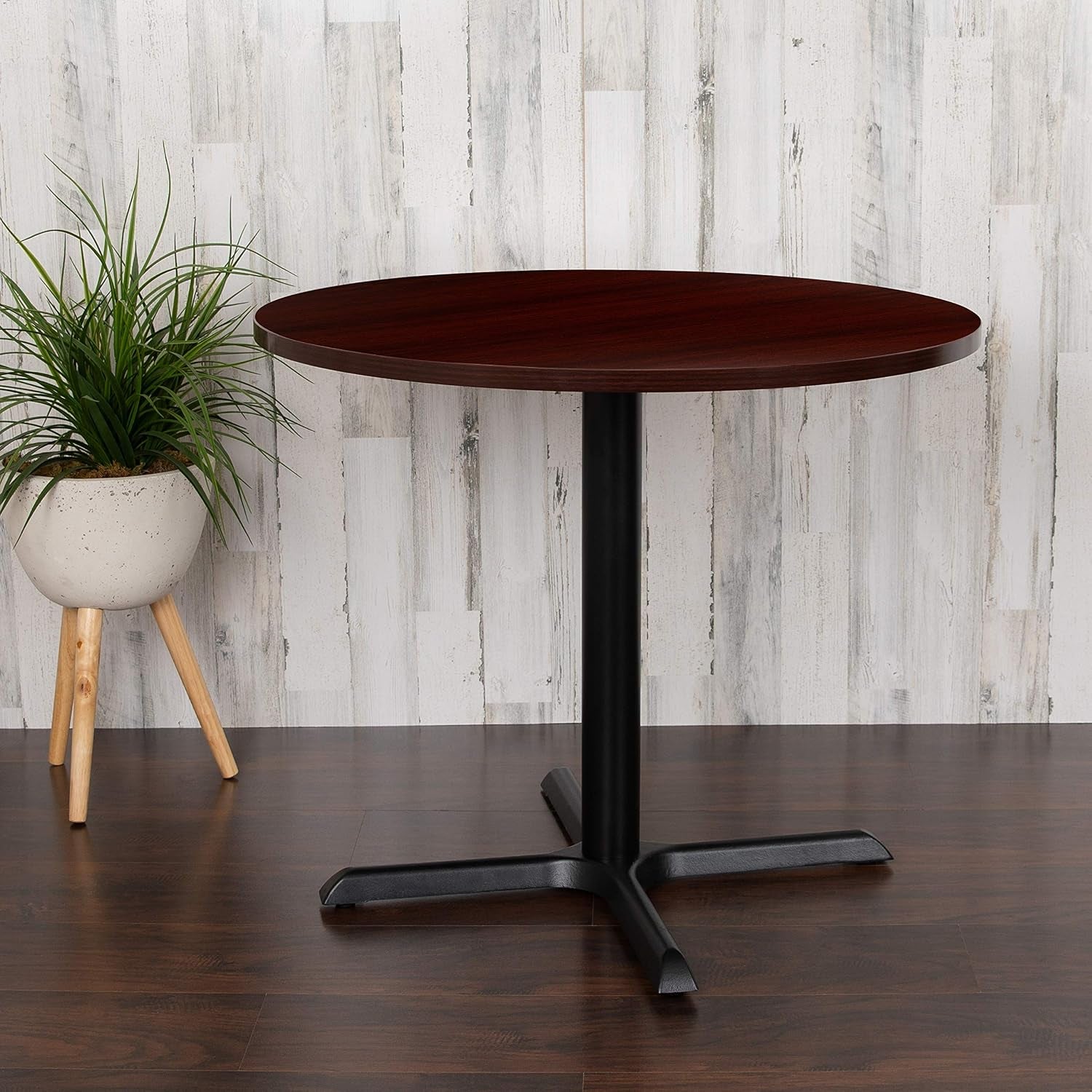 Side view of round mahogany tabletop on a black pedestal base beside a potted plant in a staged setting.