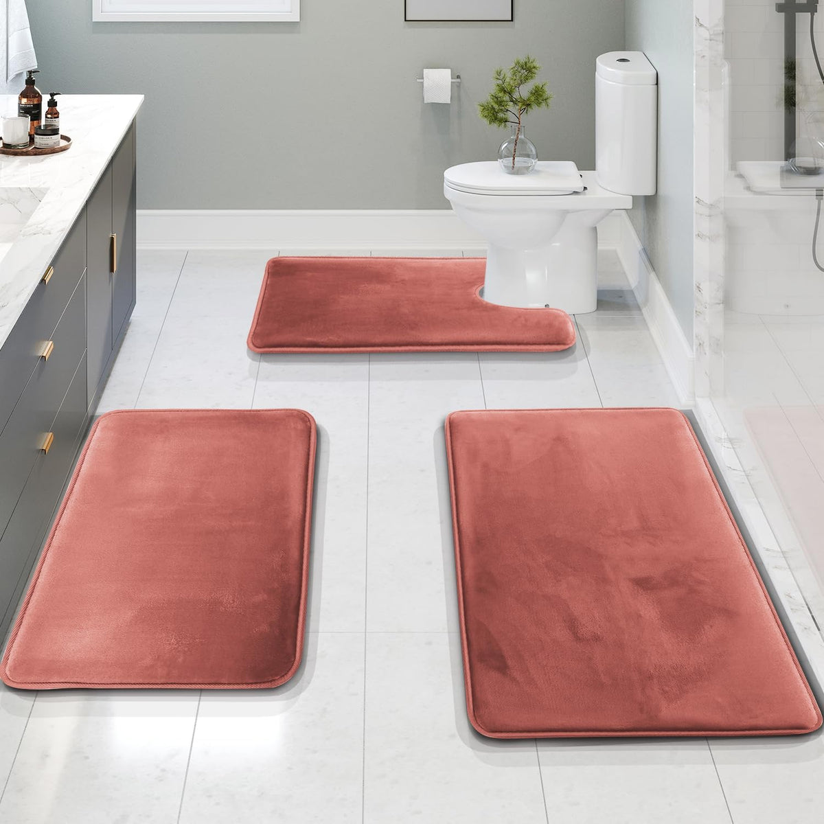 Salmon‑colored three-piece bath rug set arranged on a white tiled bathroom floor around a toilet and vanity.