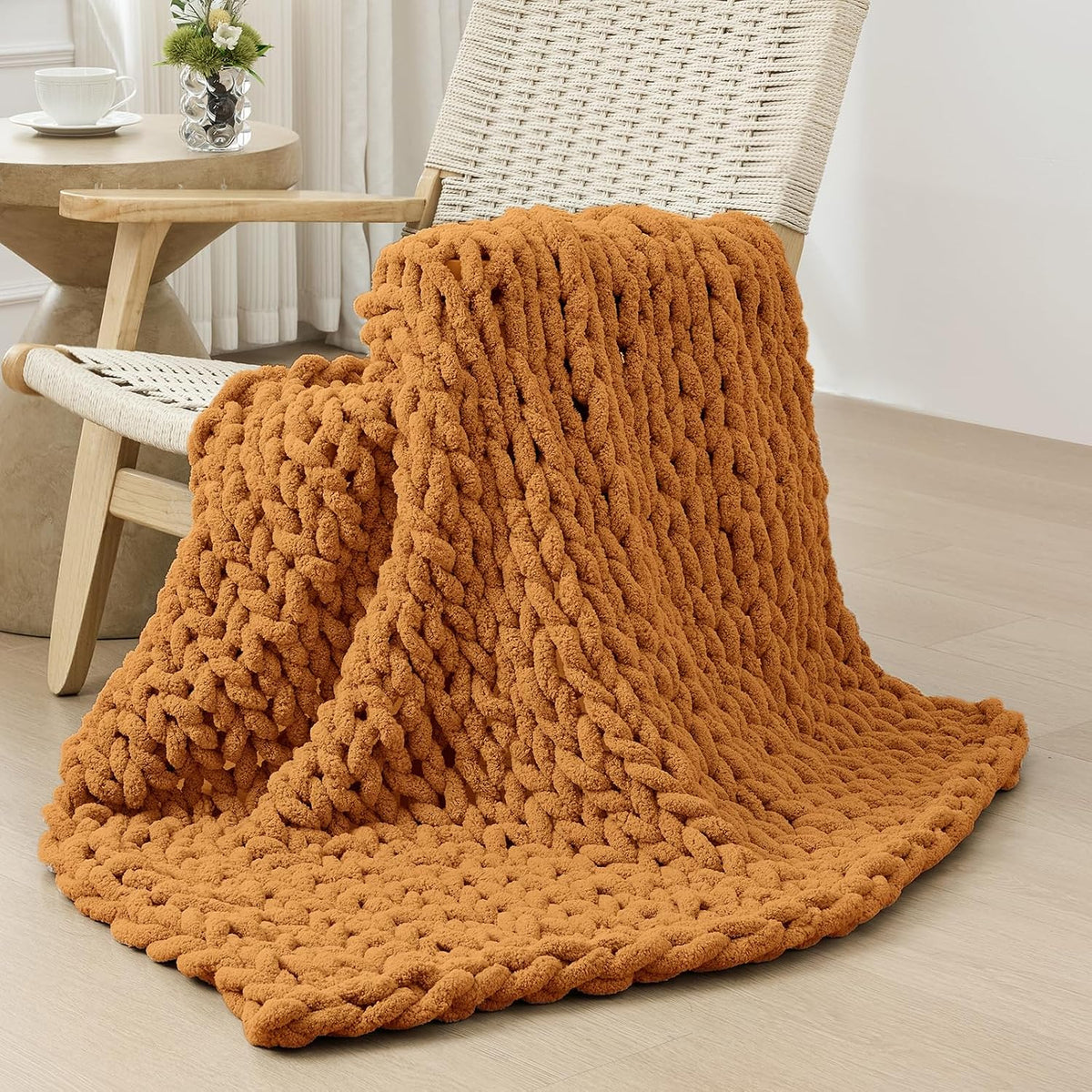 Burnt orange chunky knit throw casually draped over a woven wood chair beside a side table.