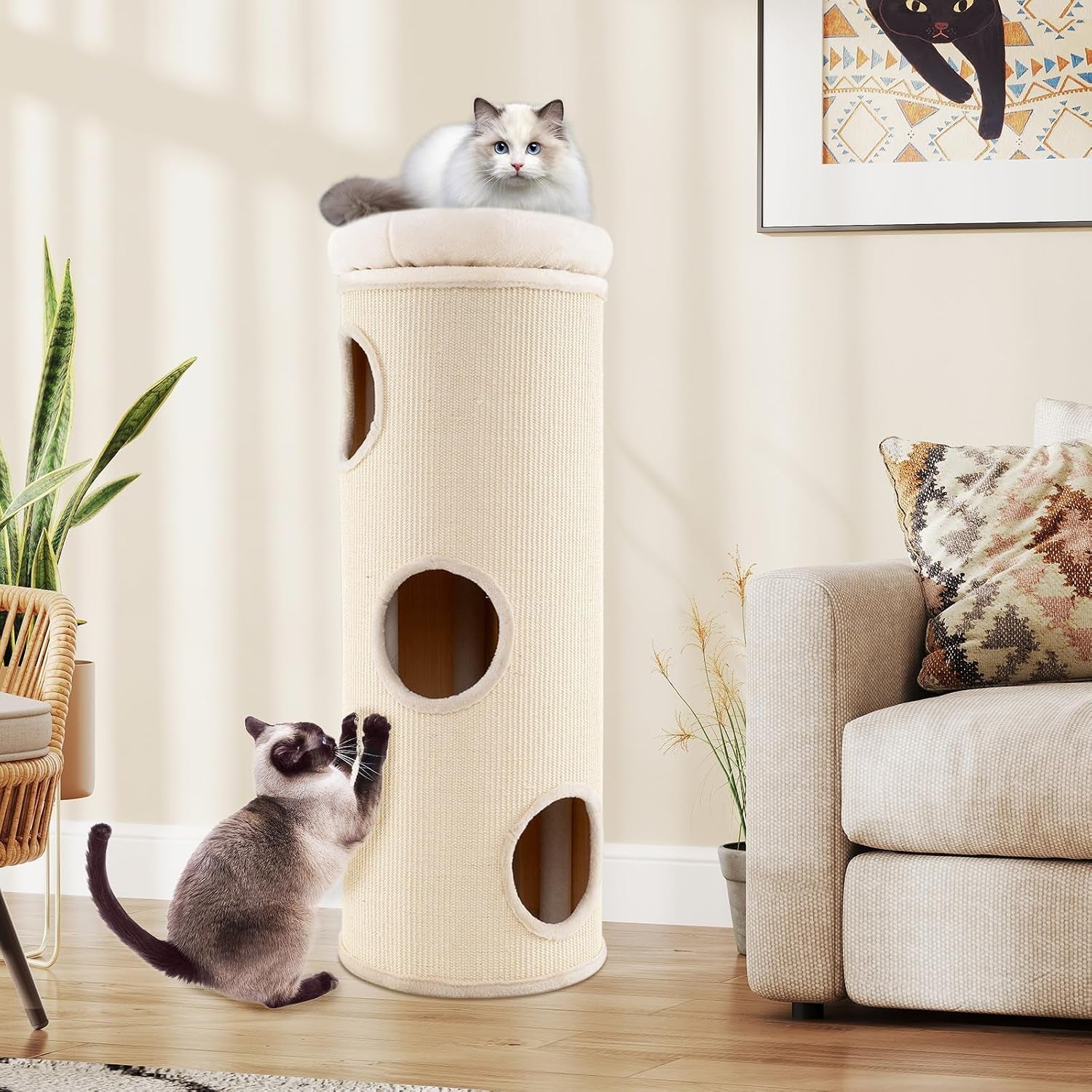 Living room scene with a cream barrel cat condo between plants and a sofa; one cat on top perch and another scratching the