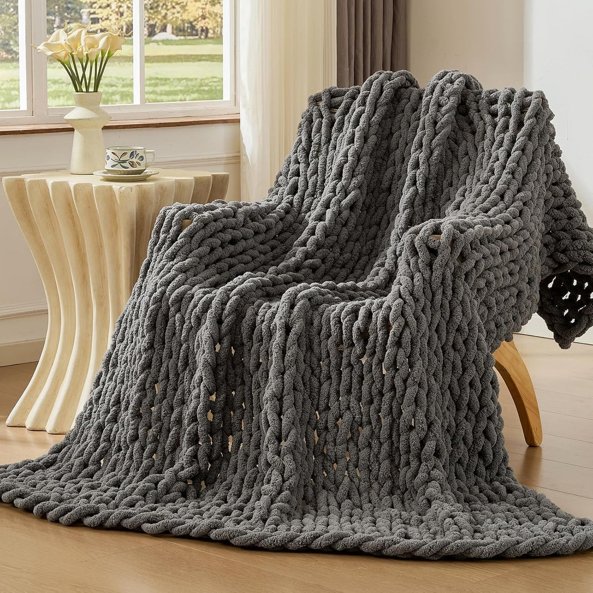 Charcoal chunky knit throw draped over a wooden lounge chair beside a sculpted side table and window.
