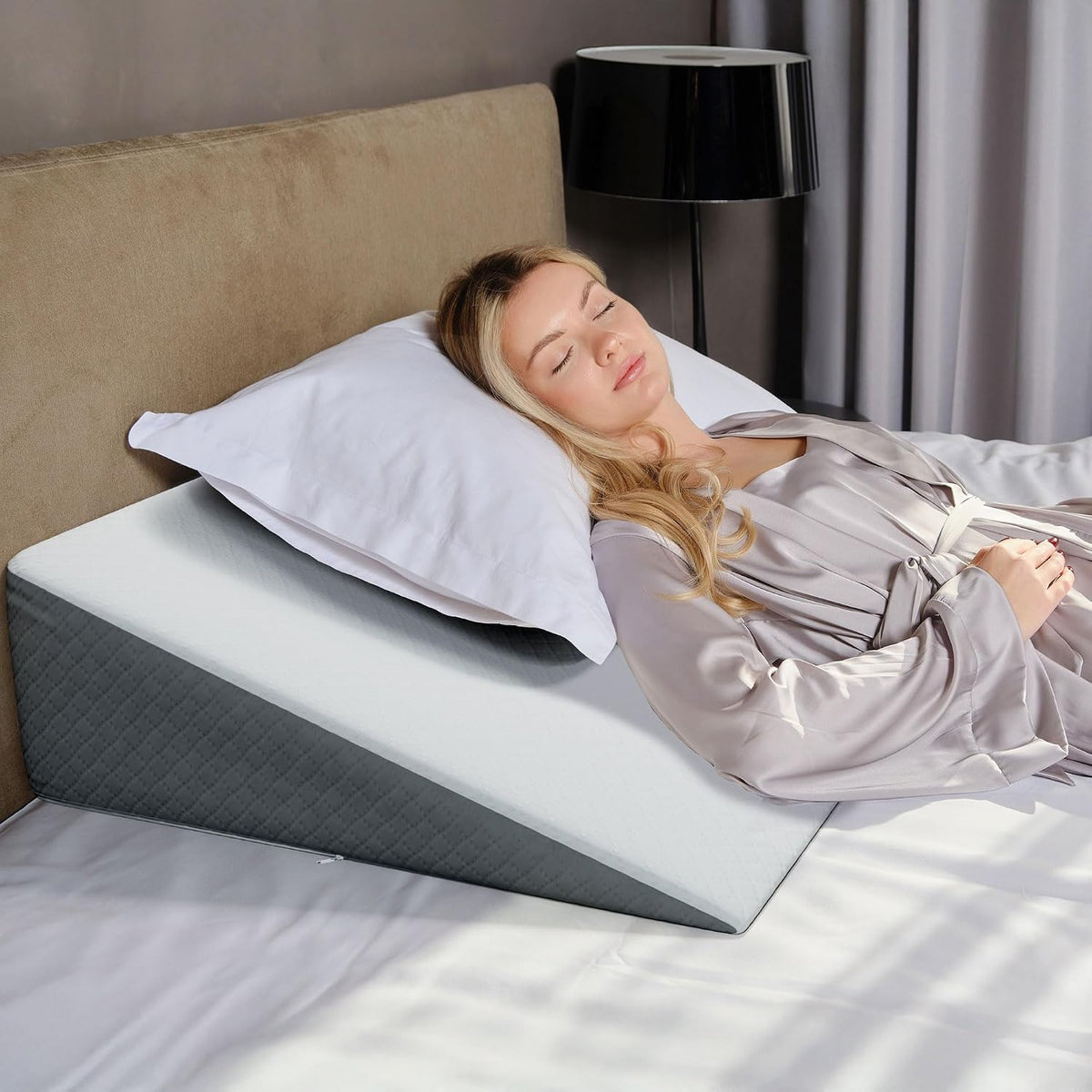 Woman lying with head on a standard pillow placed over a gray wedge, bed headboard and black bedside lamp visible.