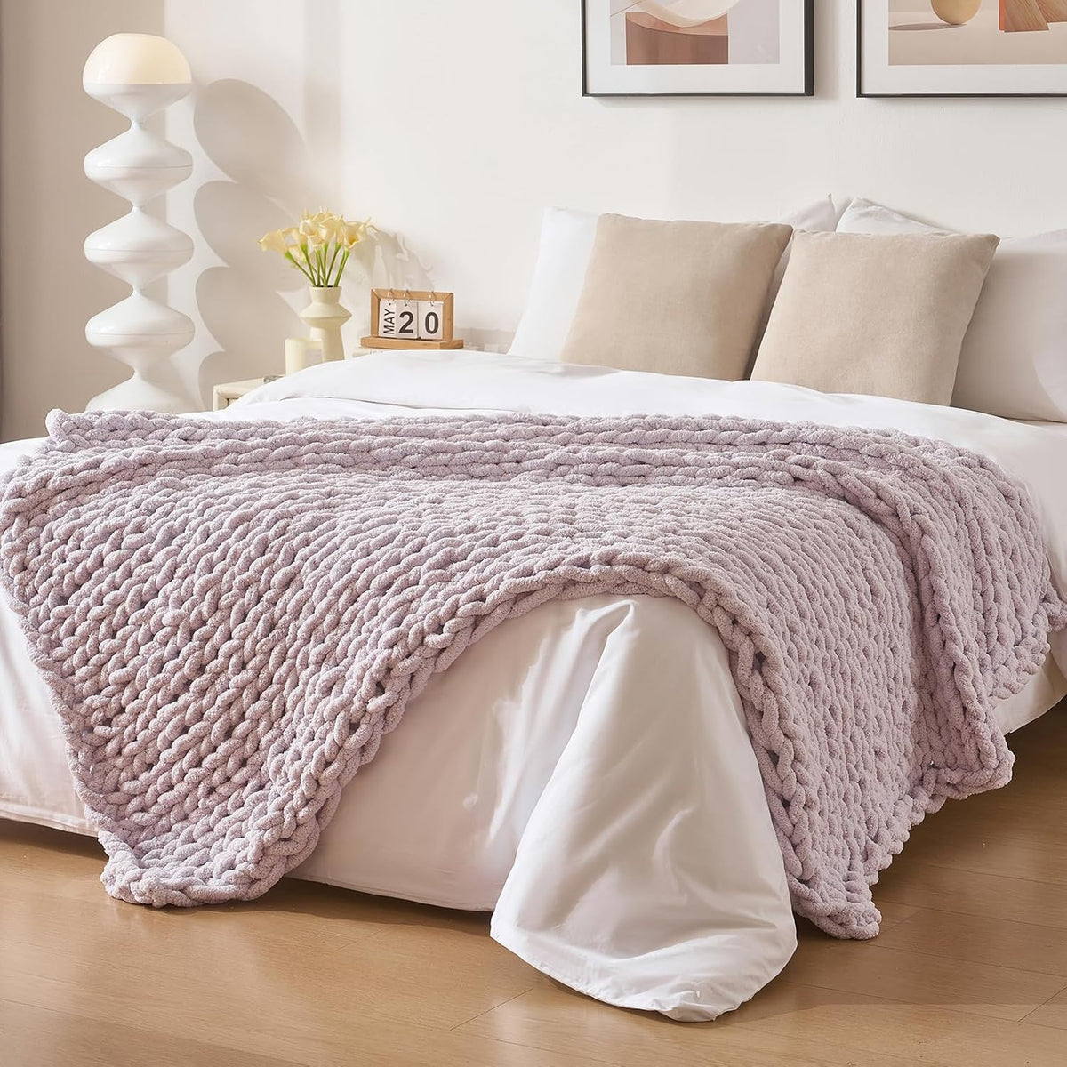 Pale lavender chunky knit throw casually spread across a white bed with beige pillows.