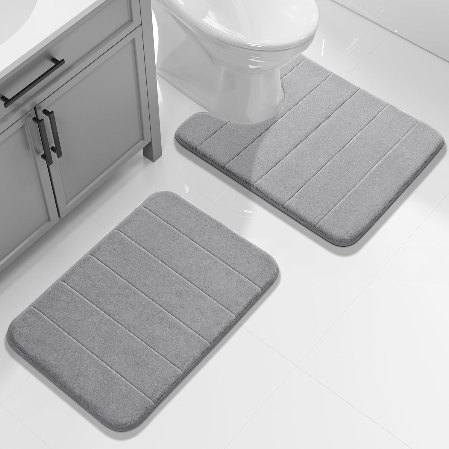 Pair of light gray bath mats placed beside a sink cabinet and in front of a toilet on glossy tile