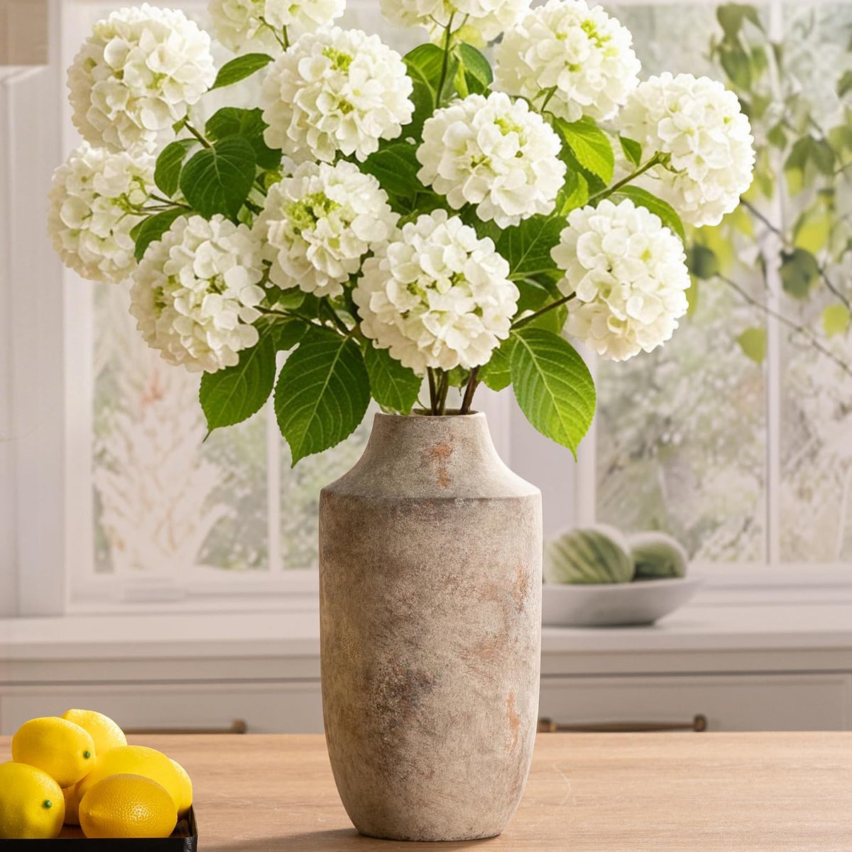 Tall weathered vase holding white hydrangeas on a kitchen table, a small tray of lemons nearby.