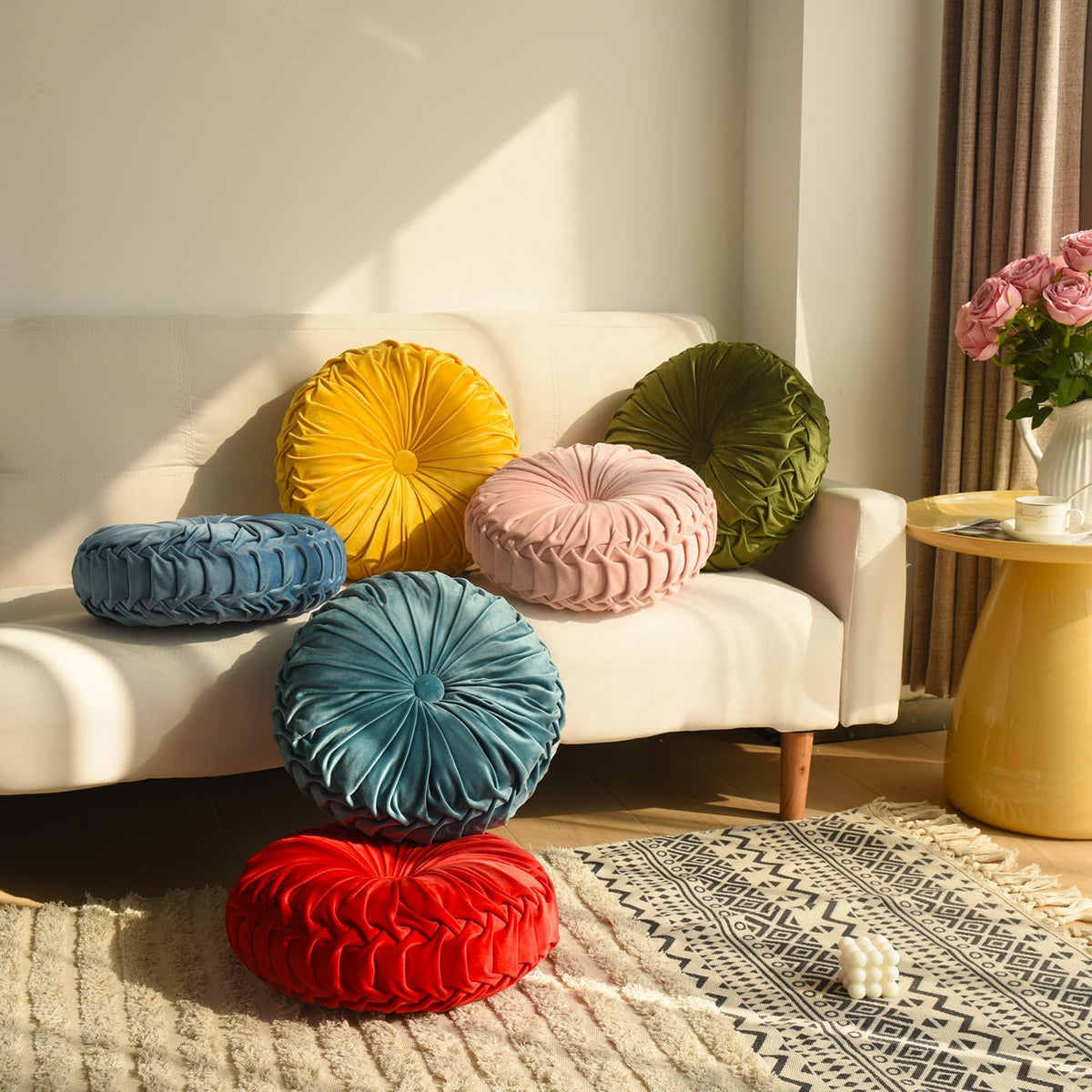 Assortment of round ruched velvet pillows in multiple colors arranged on a white sofa and the floor in a living room scene