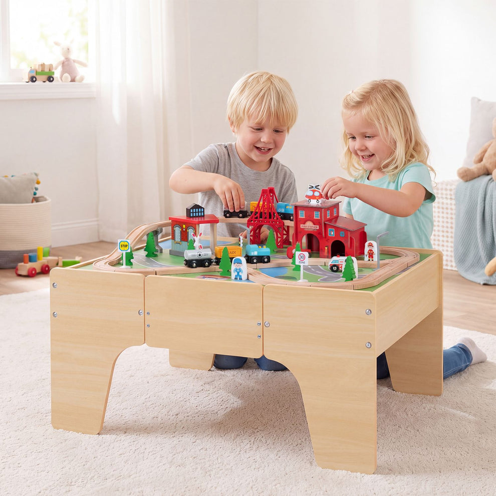 Two children play with toy trains on a wooden train table in a bright nursery.