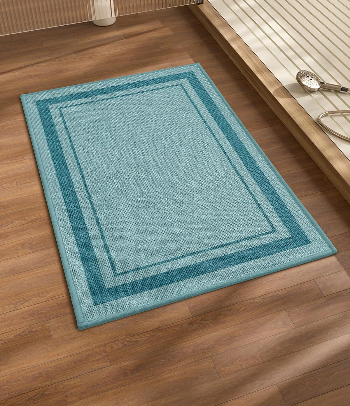 Light blue rectangular bath mat with darker double border on wood floor near a bathtub edge