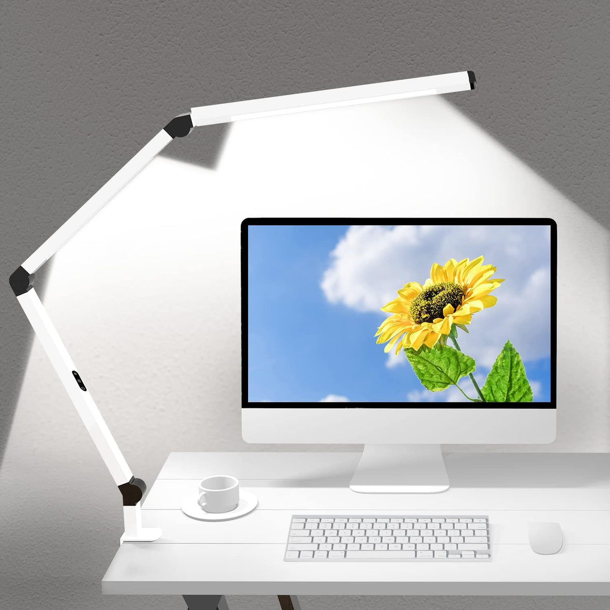 White clamp lamp angled to cast a wide beam on the wall above a monitor displaying a sunflower.