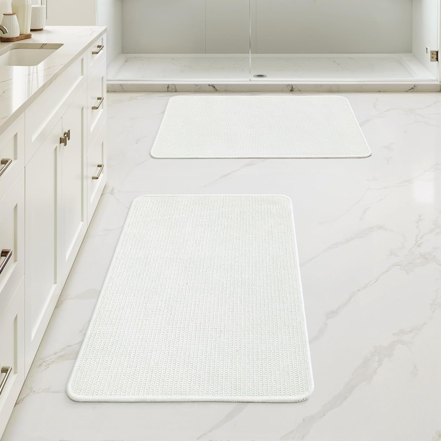 Two rectangular white bath mats arranged down a bathroom floor near a white vanity and shower.
