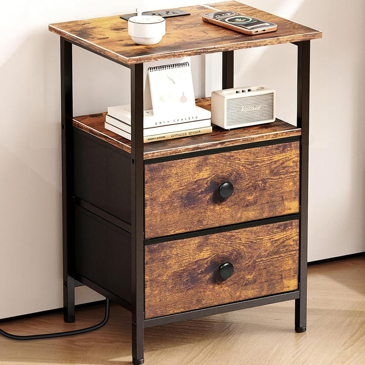 Front three-quarter view of a brown wood-finish nightstand with black metal frame, two fabric drawers and shelf with books