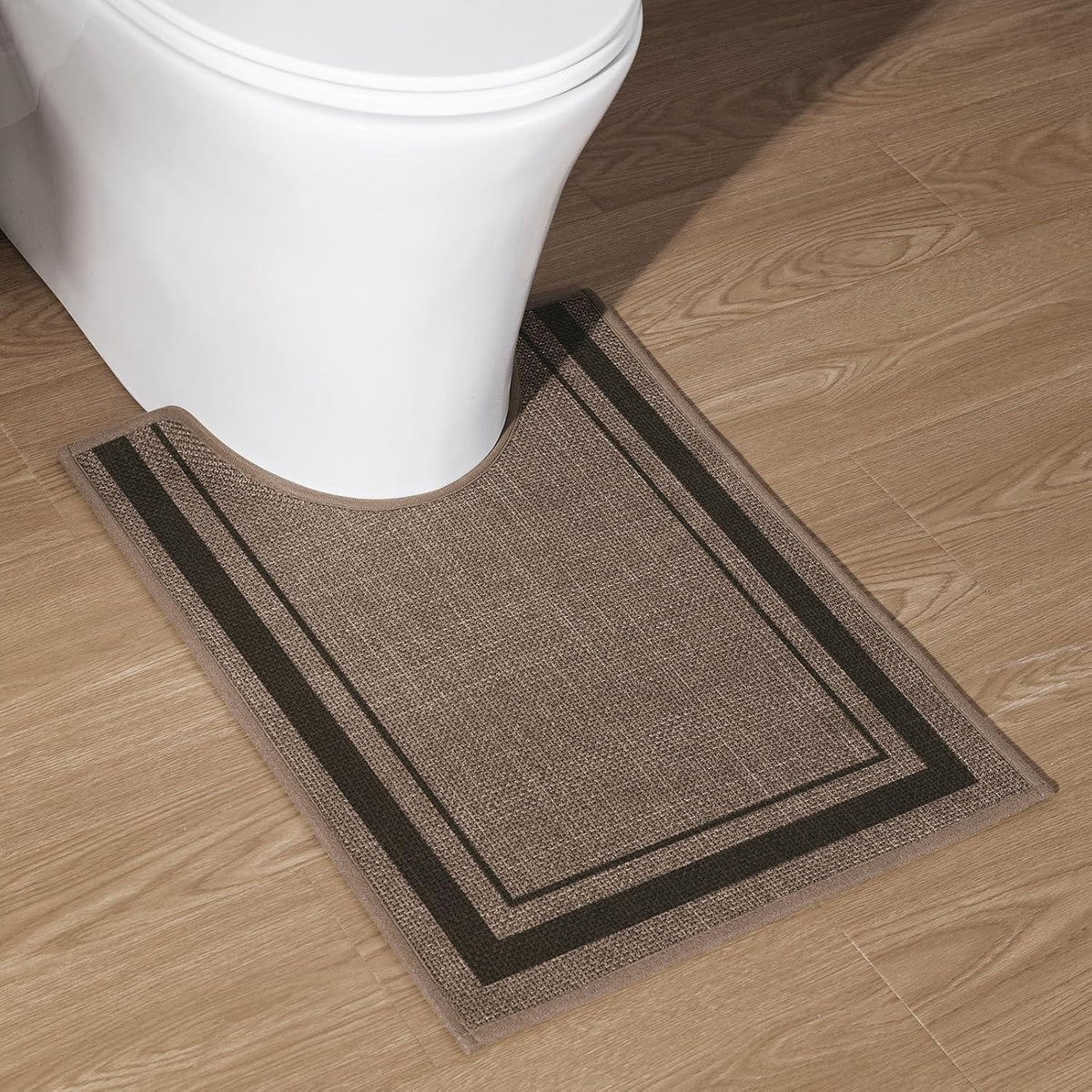 U-shaped dark beige bath rug with double dark border placed around the base of a white toilet on wood floor.