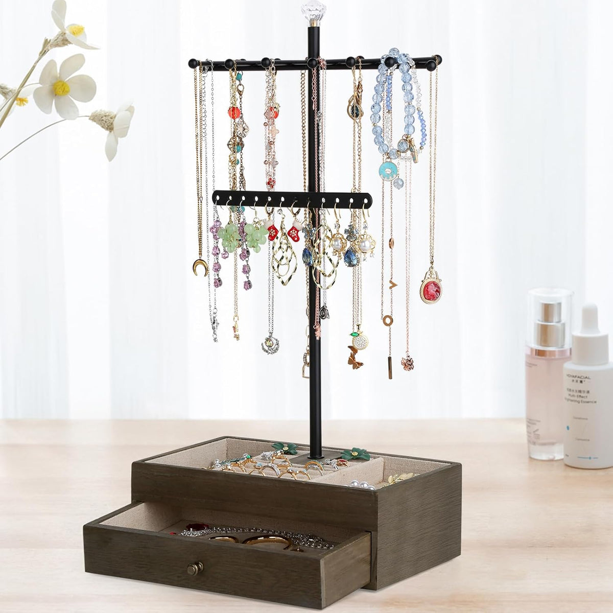 Straight-on view of the necklace tree on a darker wood base placed on a tabletop with bottles and decorative flowers in the