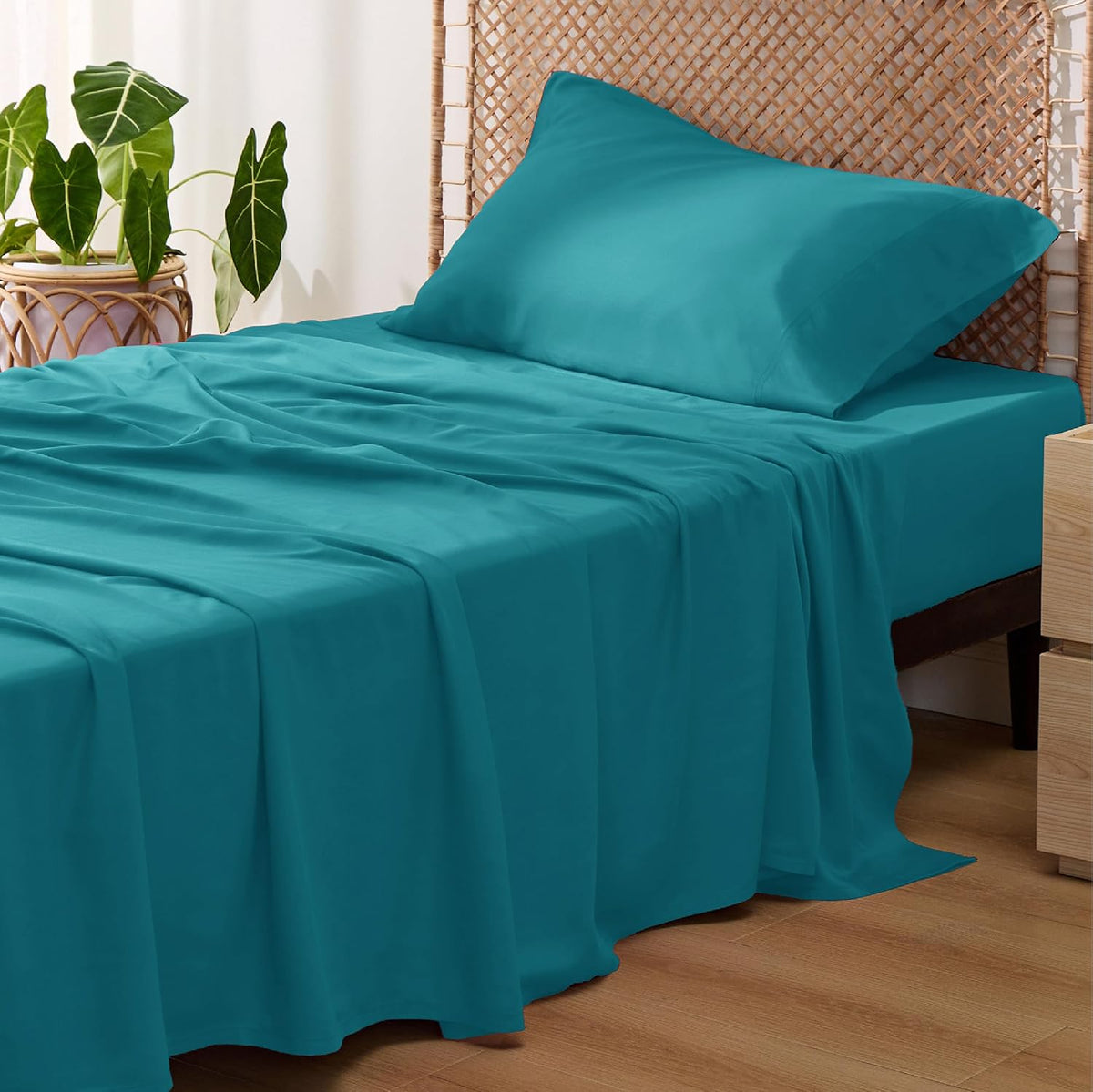 Side view of a bed dressed in teal sheets with one matching pillow against a woven headboard and a potted plant nearby.
