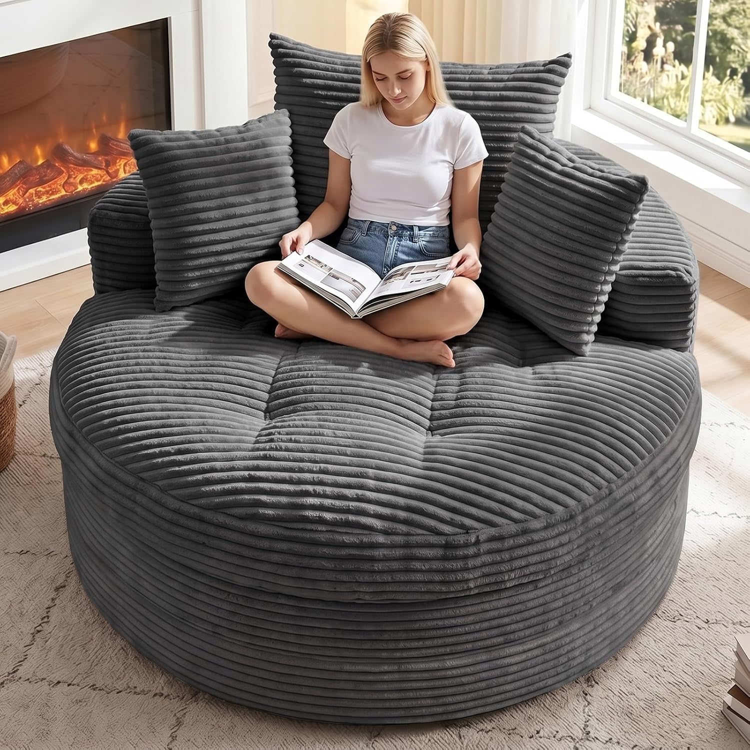 Main room shot of an oversized round gray corduroy floor chaise with three matching pillows and a woman sitting cross‑legged