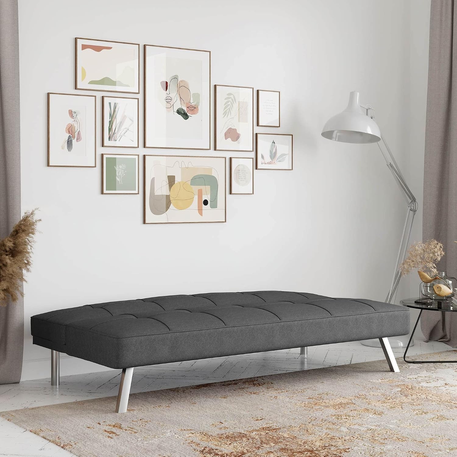 Low-profile dark gray tufted futon in a staged living room with chrome legs and gallery wall behind