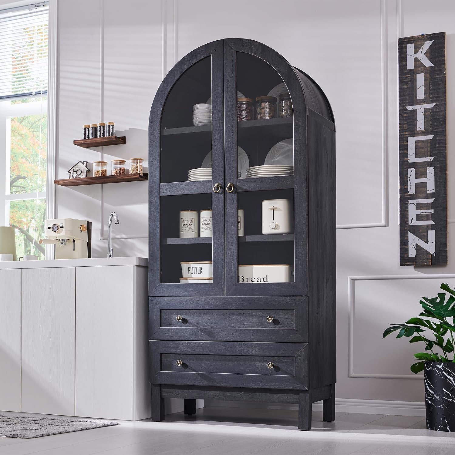 Tall arched dark wood pantry with glass-front double doors and two drawers beside a white sink in a bright kitchen.
