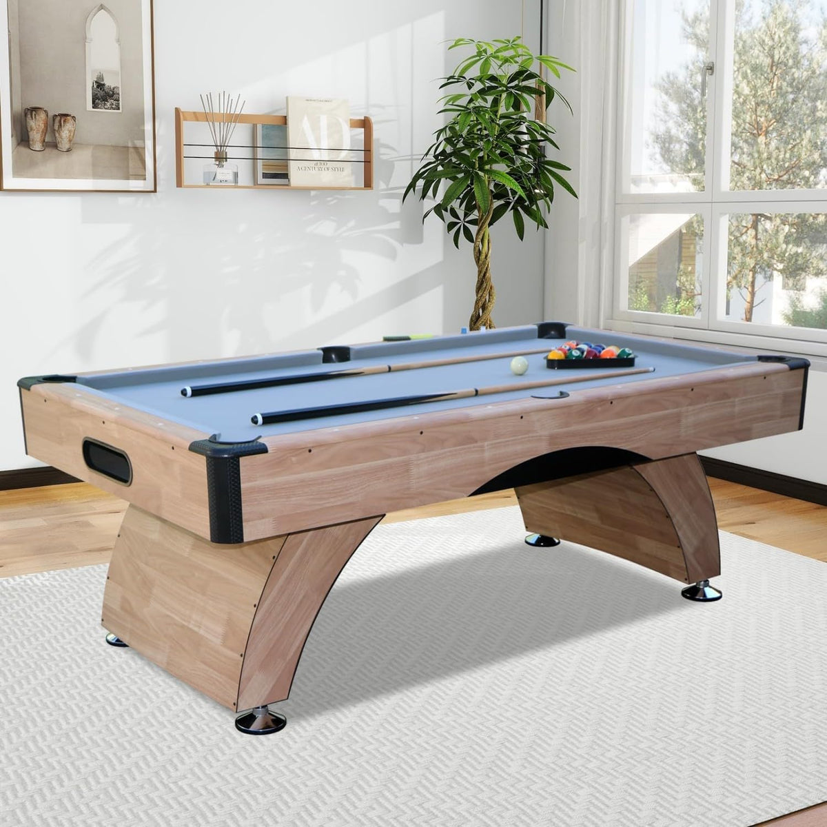 Pool table in a bright room by a window with plants and framed art, cues and racked balls on the felt.