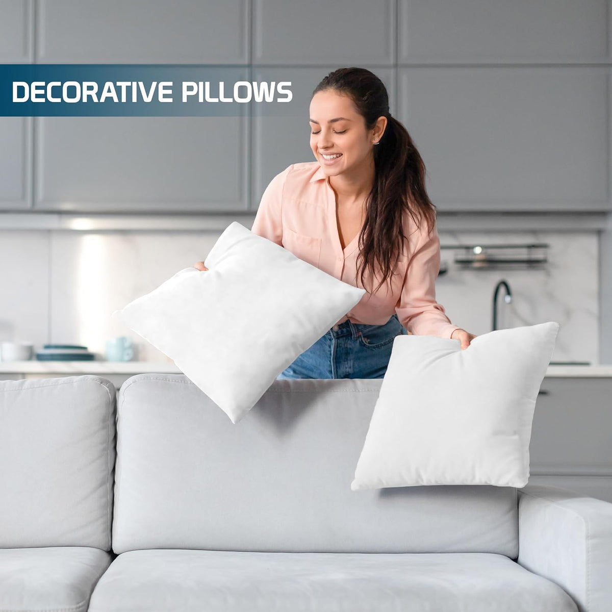 Woman placing two white throw pillow inserts on a light grey sofa in a kitchen-lifestyle background