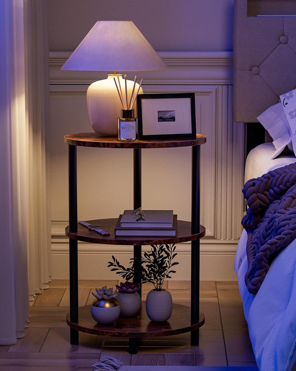 Nighttime bedroom scene of the three-tier round end table in a rustic finish used as a bedside table with a lamp and decor
