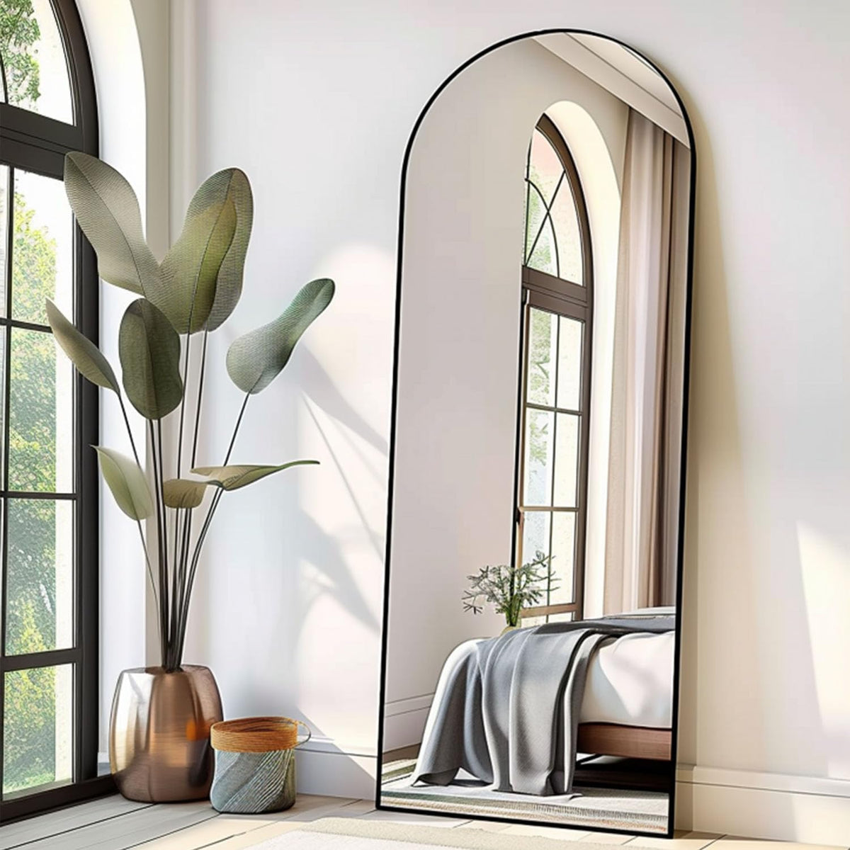 Side view of the arched mirror with black frame reflecting a bed and window, next to a tall plant.