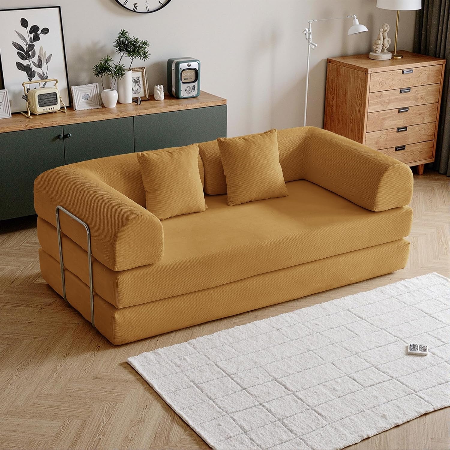 Angled view of a mustard yellow corduroy foldable sofa with three pillows and metal side rails in a cozy room.