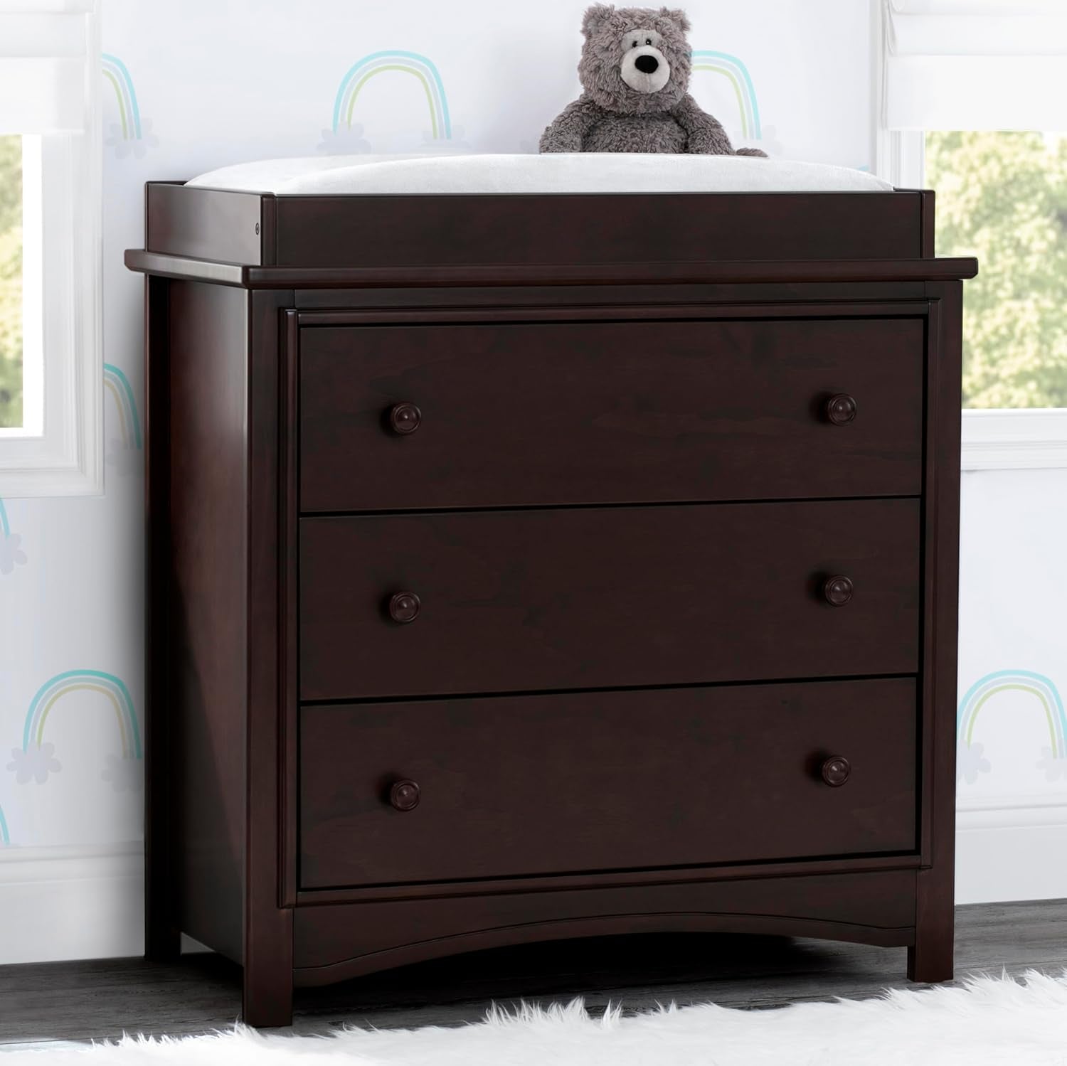 Dresser in a nursery topped with a white changing pad and a gray teddy bear, rainbow wall decals behind it.