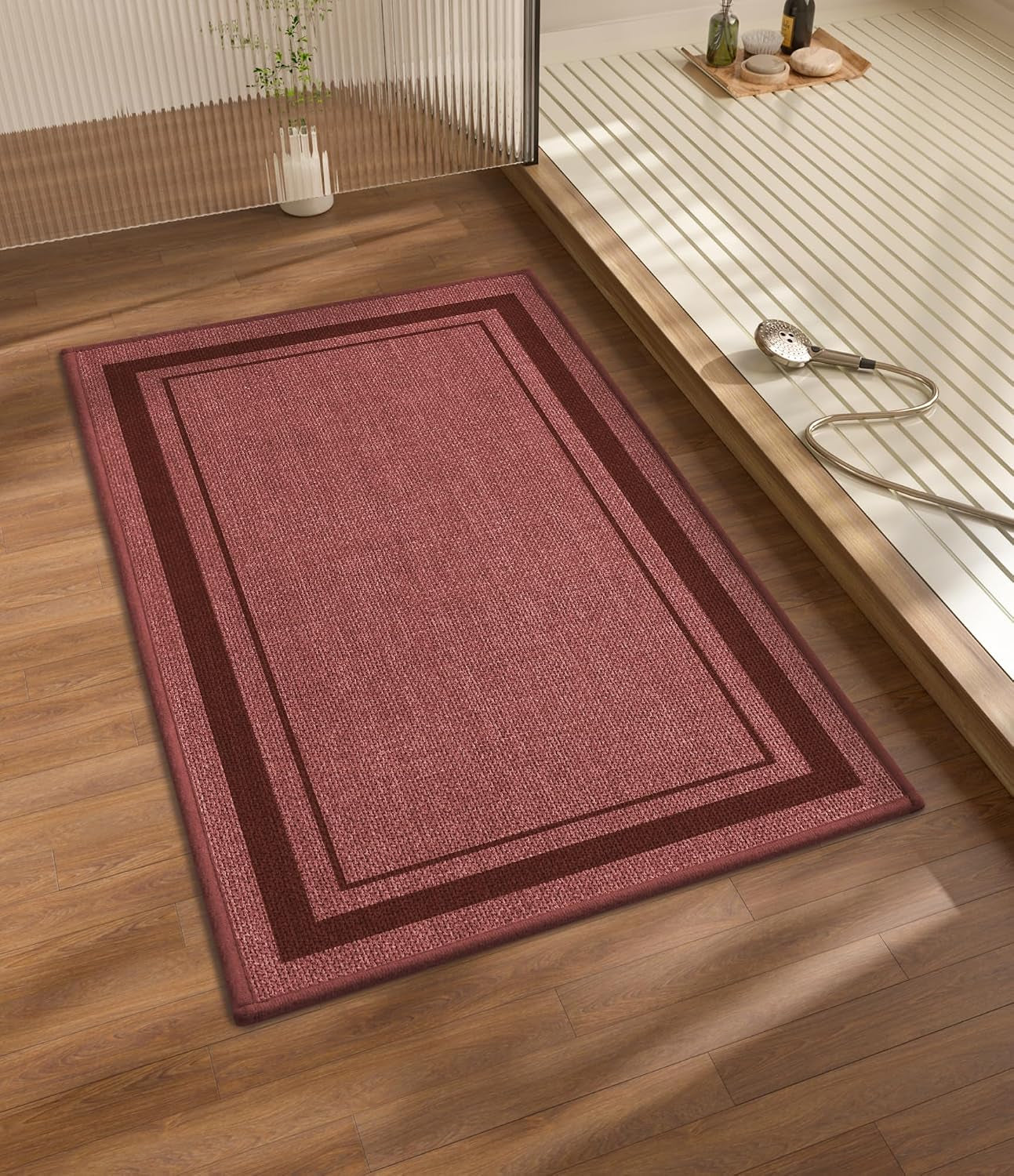 Dusty rose rectangular bath mat with two-tone border placed on wood floor near a shower area