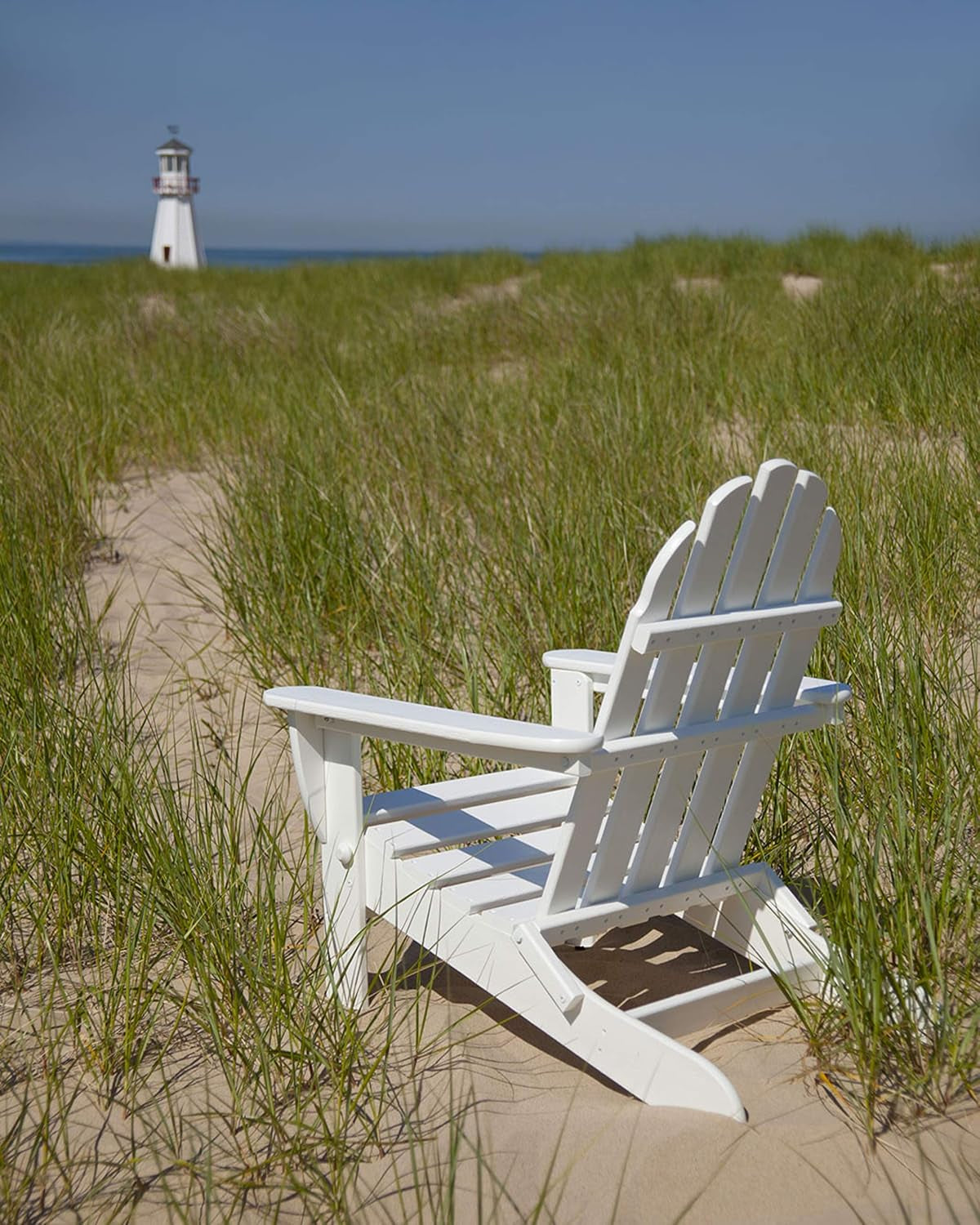 White folding Adirondack chair positioned on sandy beach among dune grass with lighthouse in the distance