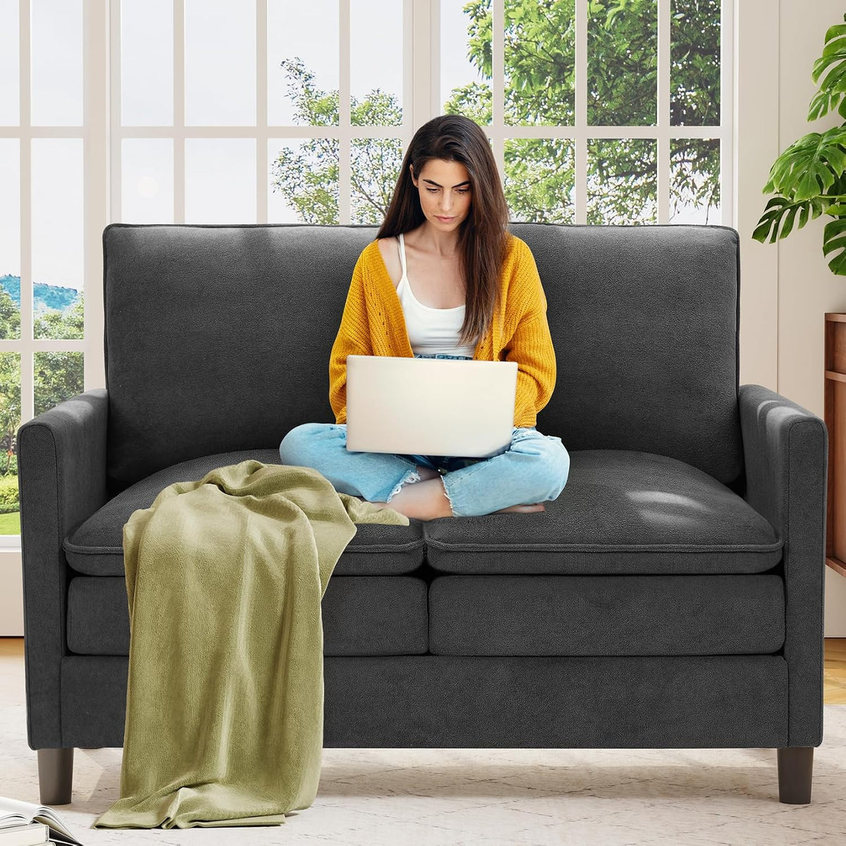Woman working on a laptop on a charcoal gray small loveseat with a tossed green blanket.
