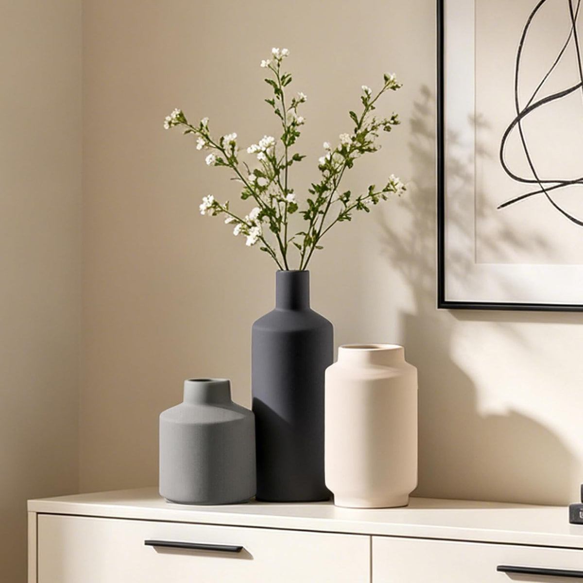 Three matte ceramic vases in cream, black and grey arranged on a console table with lamp, books and clock