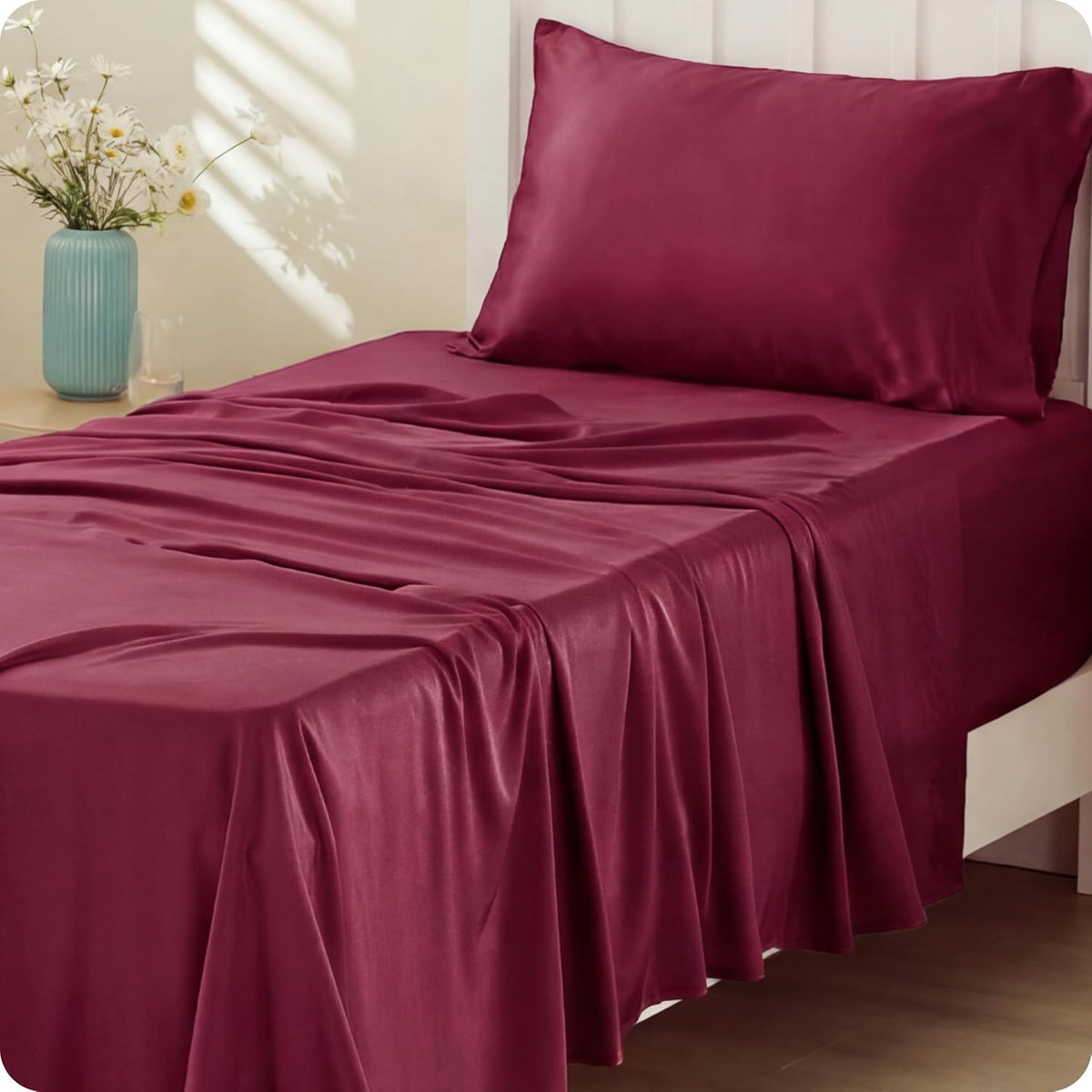 Close three-quarter view of a bed with deep burgundy sheets and pillow, fabric pooling over the bed edge onto wood floor.
